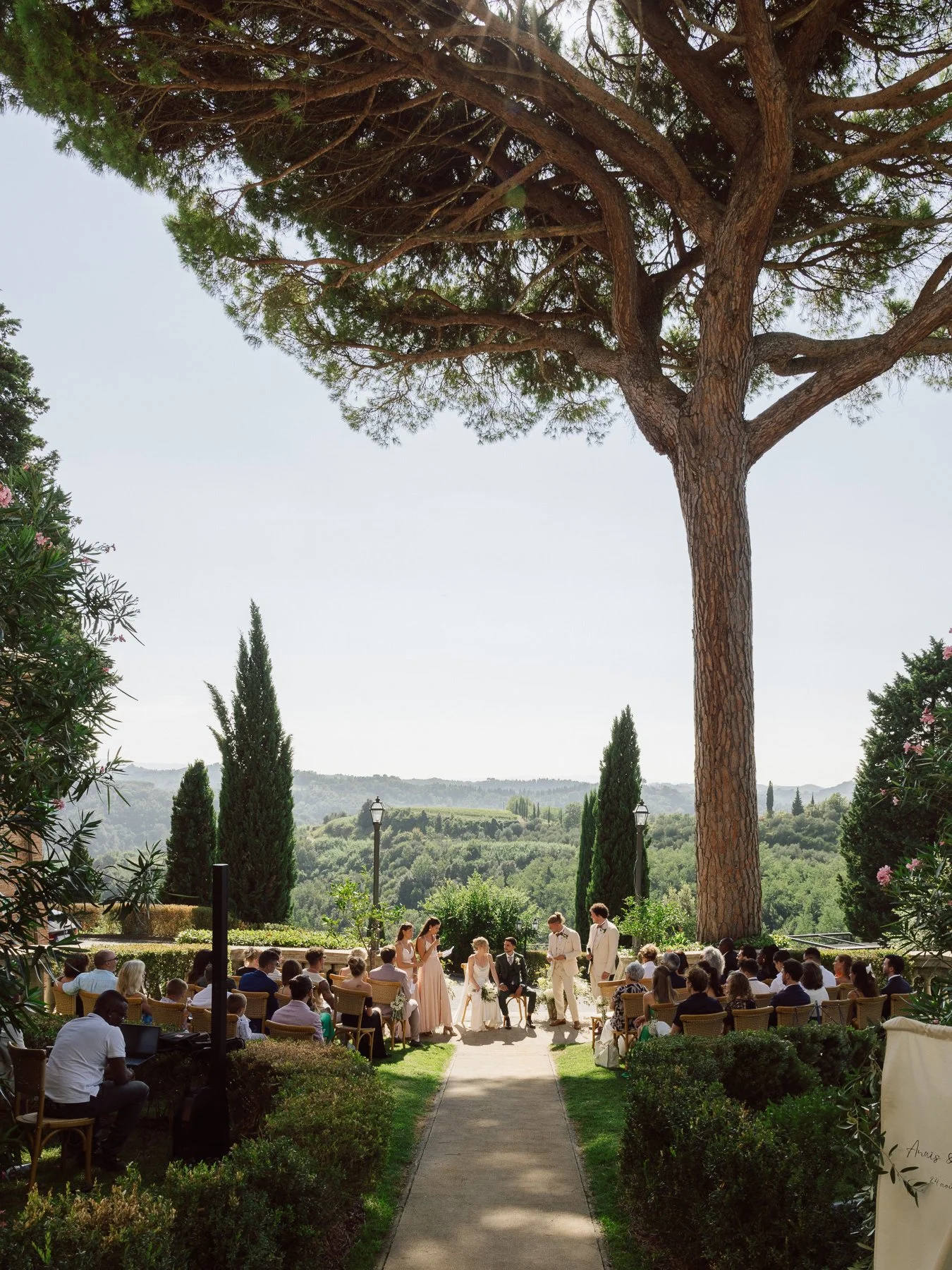 Cérémonie de mariage en plein air sous un grand arbre, avec des invités assis, des mariés et un officiant au centre, dans un jardin verdoyant avec vue sur la campagne environnante.
