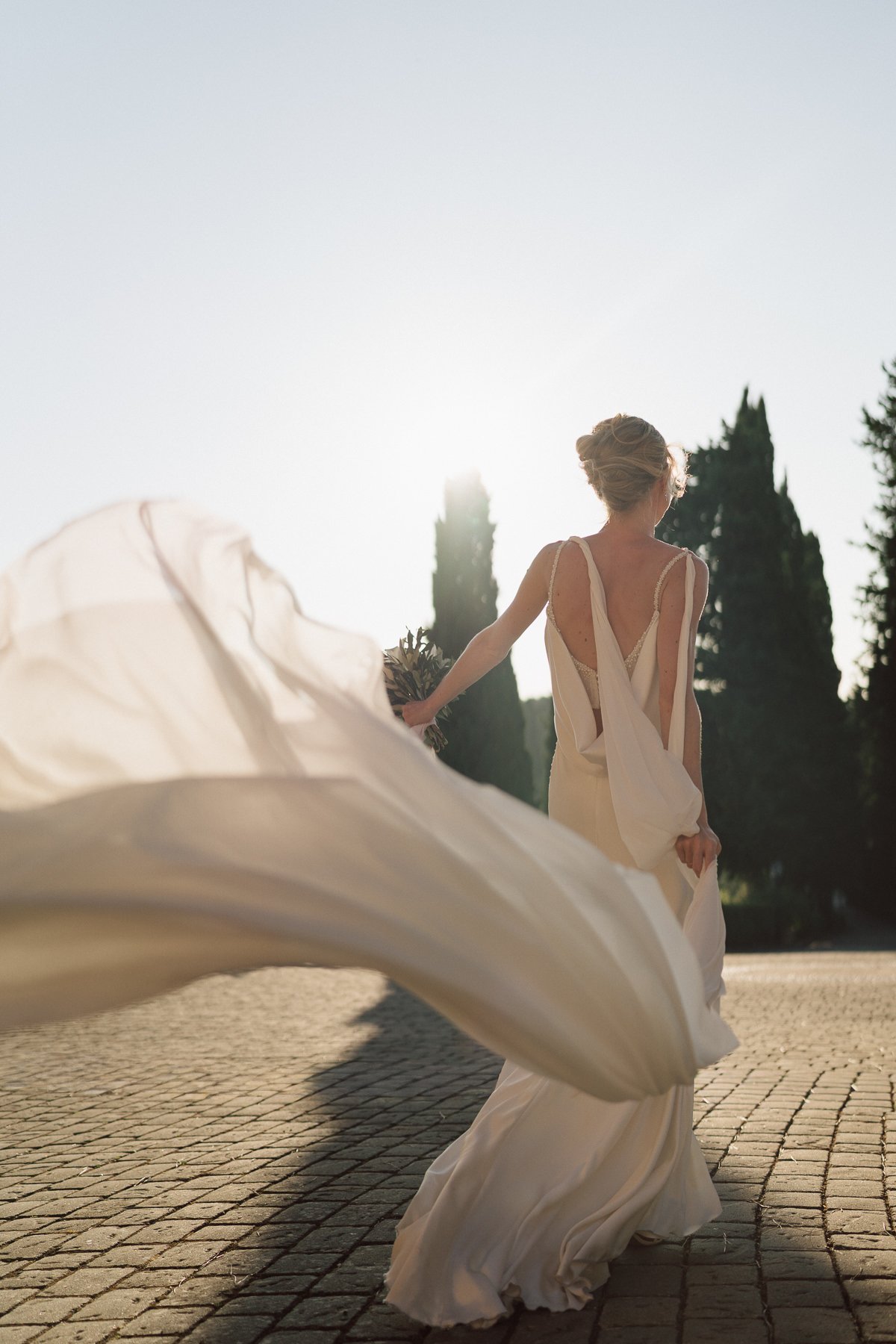 Mariée de dos en robe fluide au coucher du soleil, traîne portée par le vent, cyprès en arrière-plan dans un paysage de Toscane.