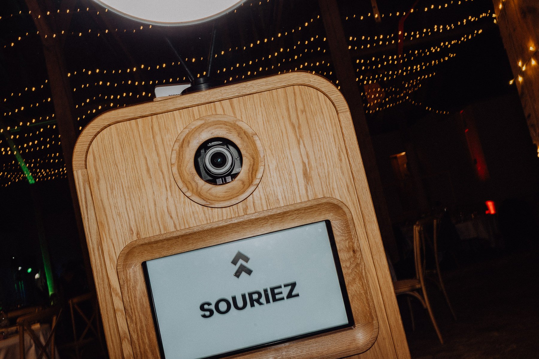 Une borne photo en bois avec une caméra en haut et un écran affichant l'inscription 'SOURIEZ' avec une flèche pointant vers le haut, dans un environnement décoré de guirlandes lumineuses.