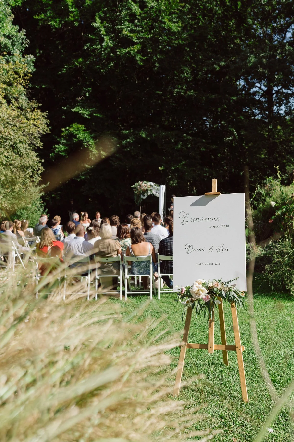 Panneau de bienvenue du mariage de Diana et Loïc lors de la cérémonie laïque en extérieur au Castel de Bois-Genoud, avec les invités en arrière-plan