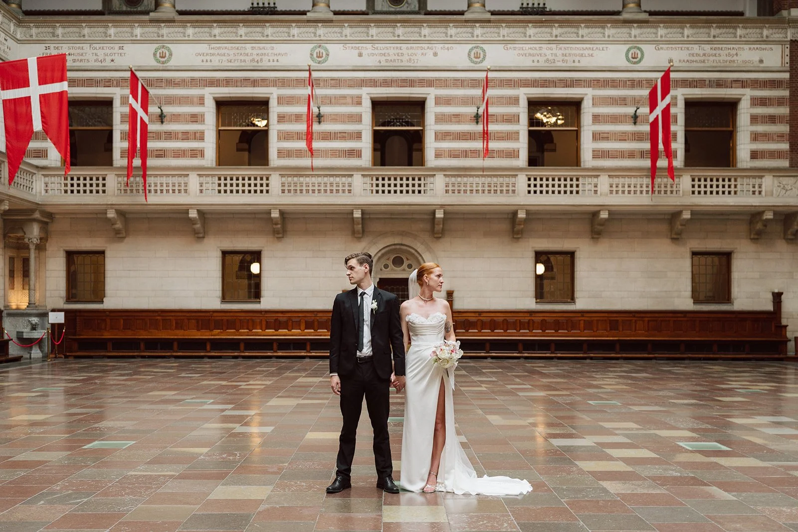 Un couple de mariés debout dans une grande salle avec des drapeaux danois accrochés au plafond. La mariée porte une robe blanche à une épaule, tenant un bouquet de fleurs, tandis que le marié porte un costume noir avec une cravate, ils tiennent la ma