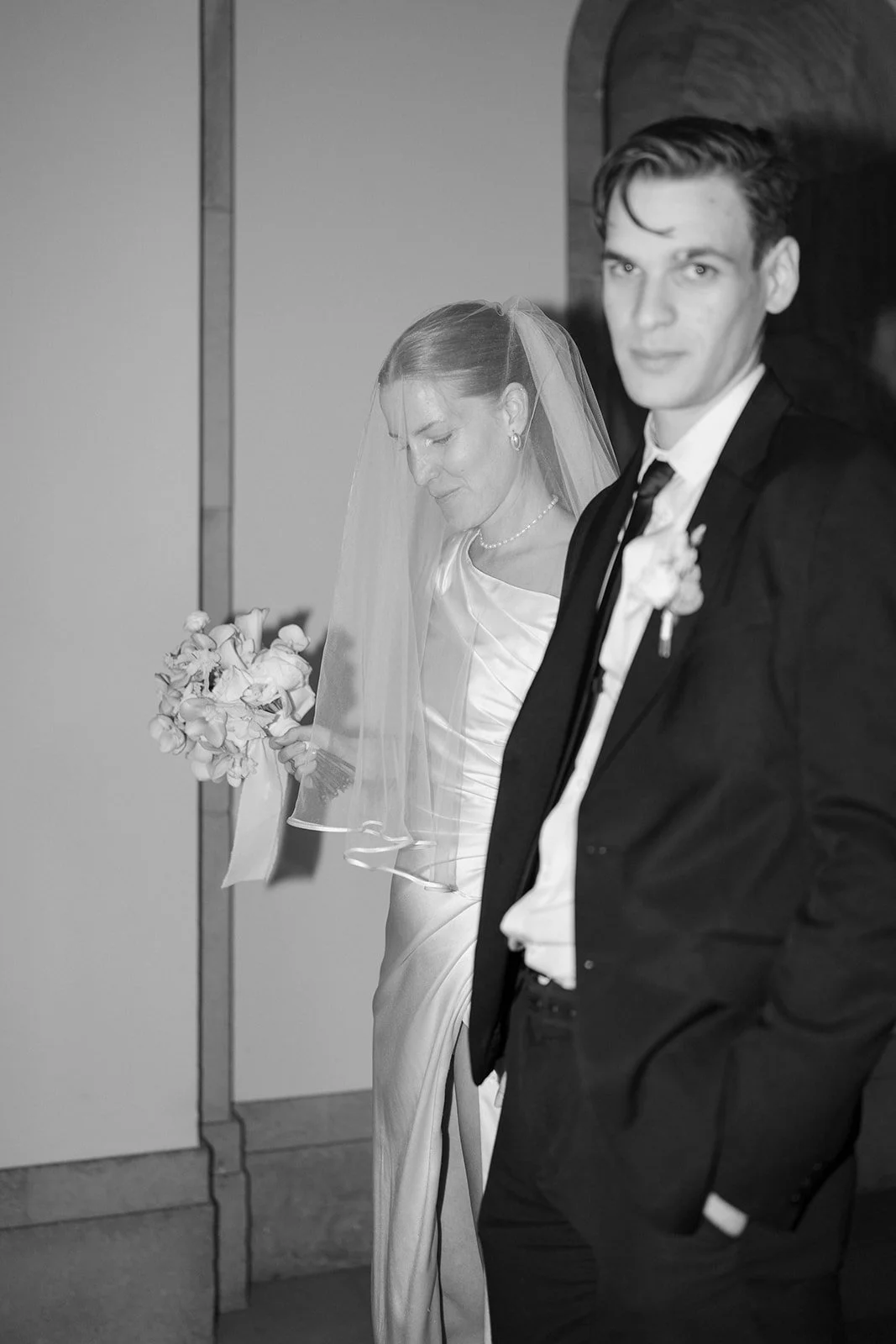 Un couple de mariés, la femme portant une robe de mariée et un voile, tient un bouquet de fleurs ; l'homme porte un costume noir avec une cravate, dans un cadre intérieur.