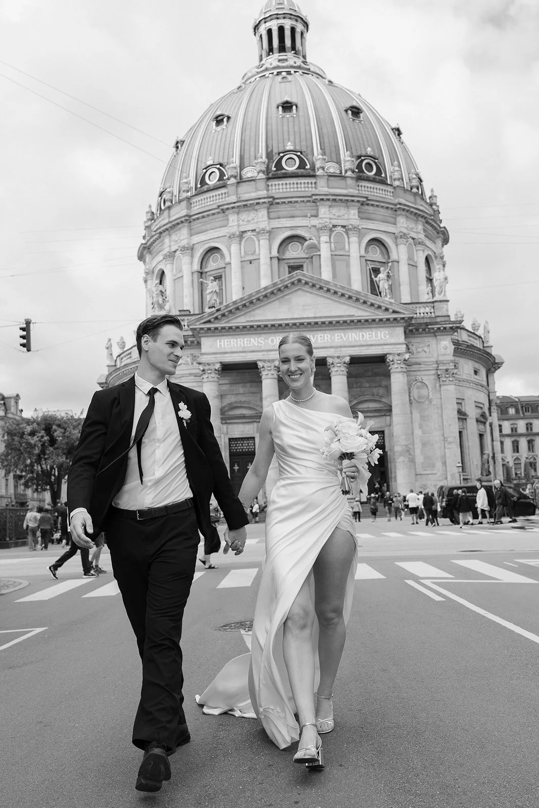Un couple en mariage, la femme tenant un bouquet, traversant une rue à Berlin avec la cathédrale Humboldt en arrière-plan