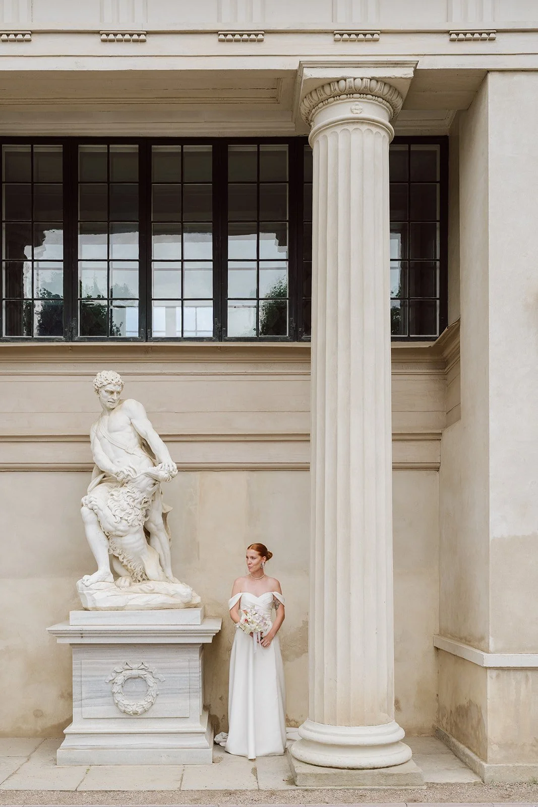 Une femme en robe blanche tenant un bouquet, se tenant près d'une statue en marbre représentant une scène mythologique, dans un bâtiment avec des colonnes et de grandes fenêtres