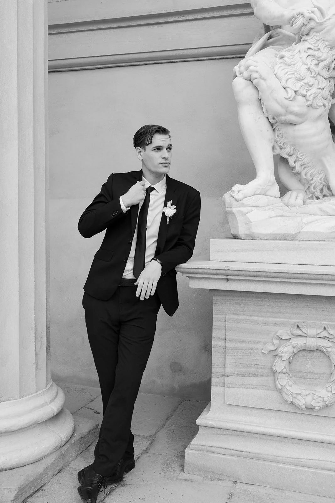 Un jeune homme en costume noir, debout, à côté d'une statue en marbre d'un lion, dans un bâtiment avec des murs en pierre.