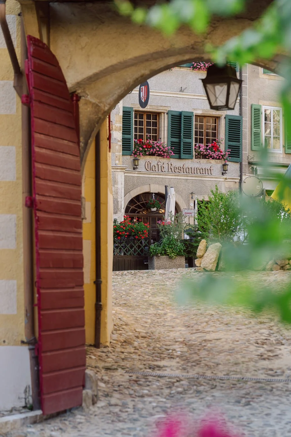 Vue sous les arcades du village de Romainmôtier, avec le café-restaurant fleuri à proximité du Jardin Baillival, dans une ambiance authentique et chaleureuse.
