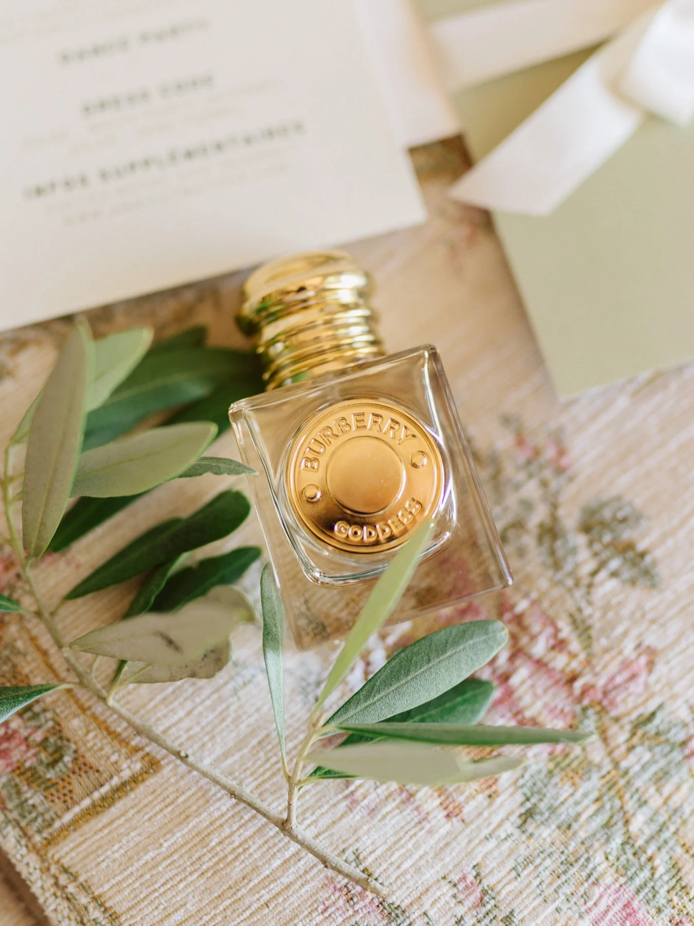 Flacon de parfum Burberry posé avec une branche d’olivier sur un faire-part de mariage aux tons vert olive, détail élégant de préparation de mariée en Toscane