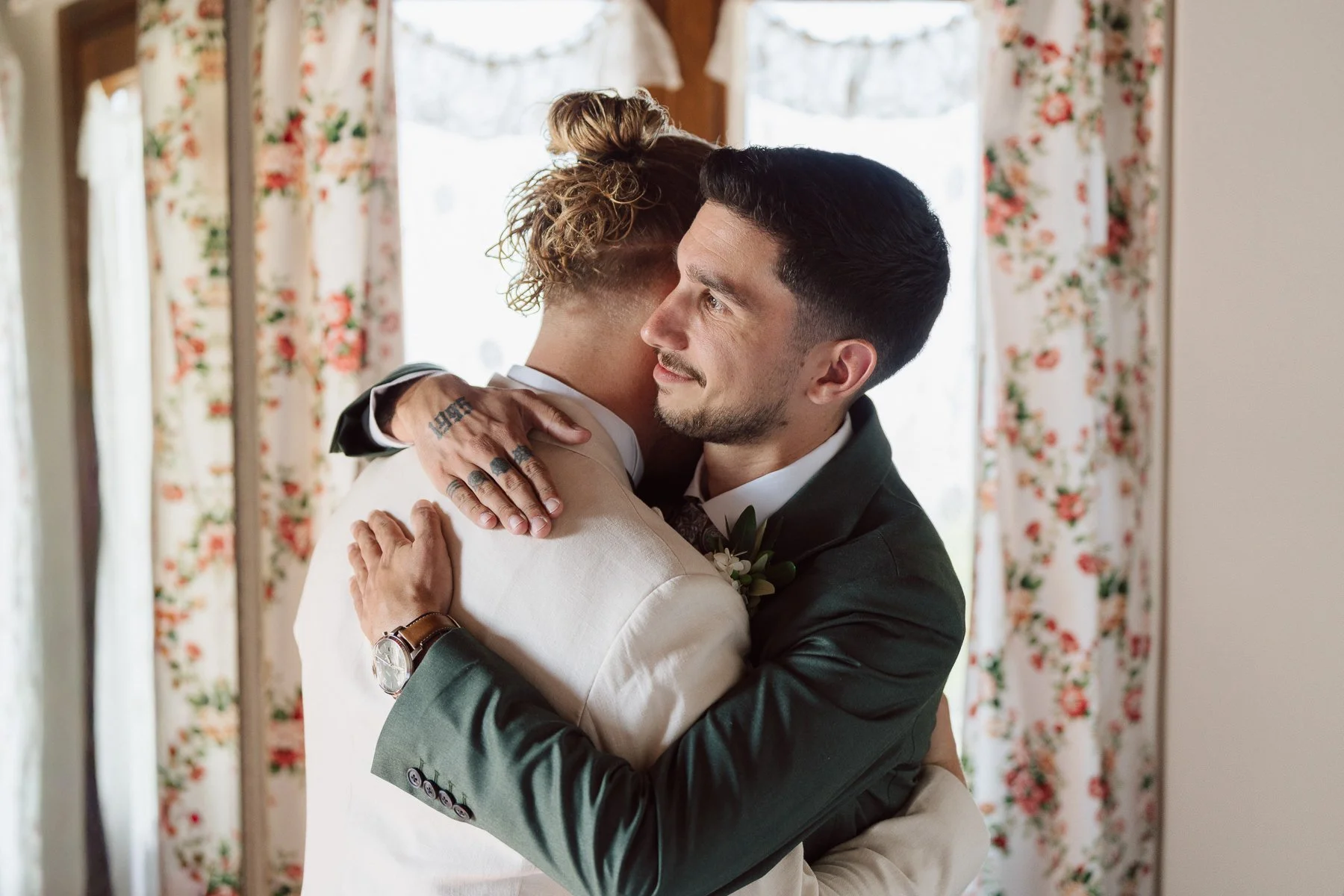 Le marié enlace un proche témoin avec émotion pendant les préparatifs du mariage en Toscane, dans une chambre lumineuse aux rideaux fleuris.