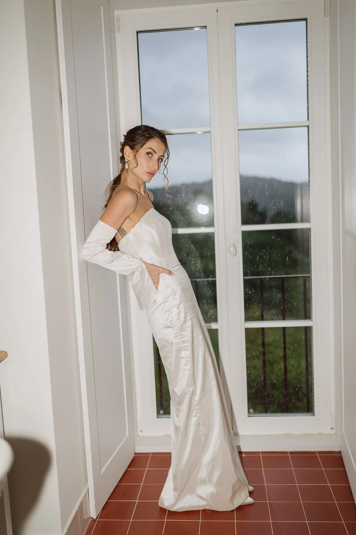 Mariée en robe satinée élégante posant près d’une fenêtre pendant les préparatifs de mariage, ambiance éditoriale chic et lumineuse