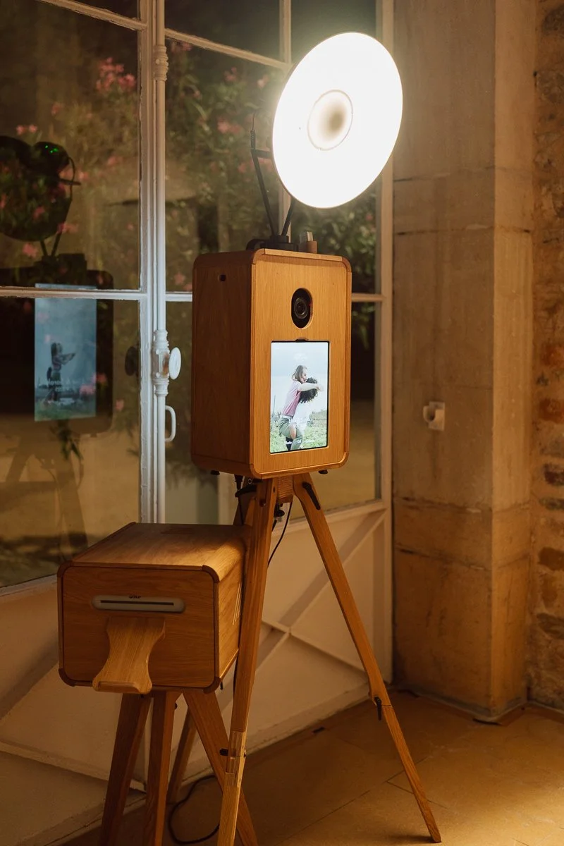 Une cabine photo en bois avec un écran, située sur un trépied, avec une lumière bright à l'arrière et une fenêtre en arrière-plan, dans une pièce en pierre.