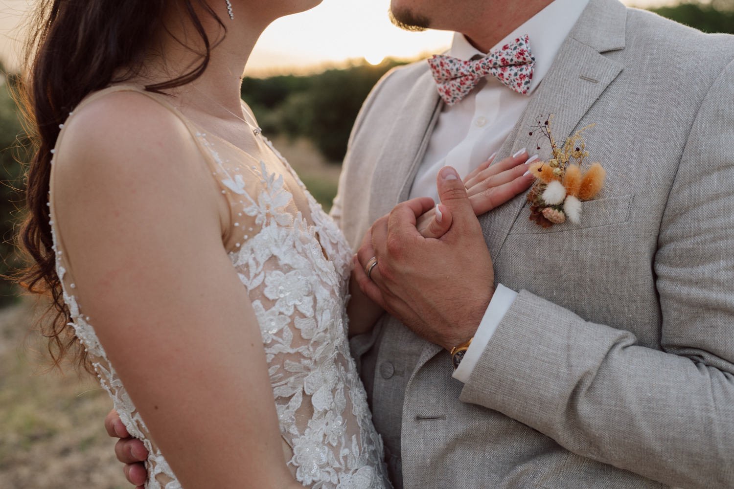 Gros plan sur les mains des mariés lors d’un moment tendre, la mariée en robe en dentelle et le marié en costume beige avec nœud papillon fleuri et boutonnière séchée, au coucher de soleil en Provence.