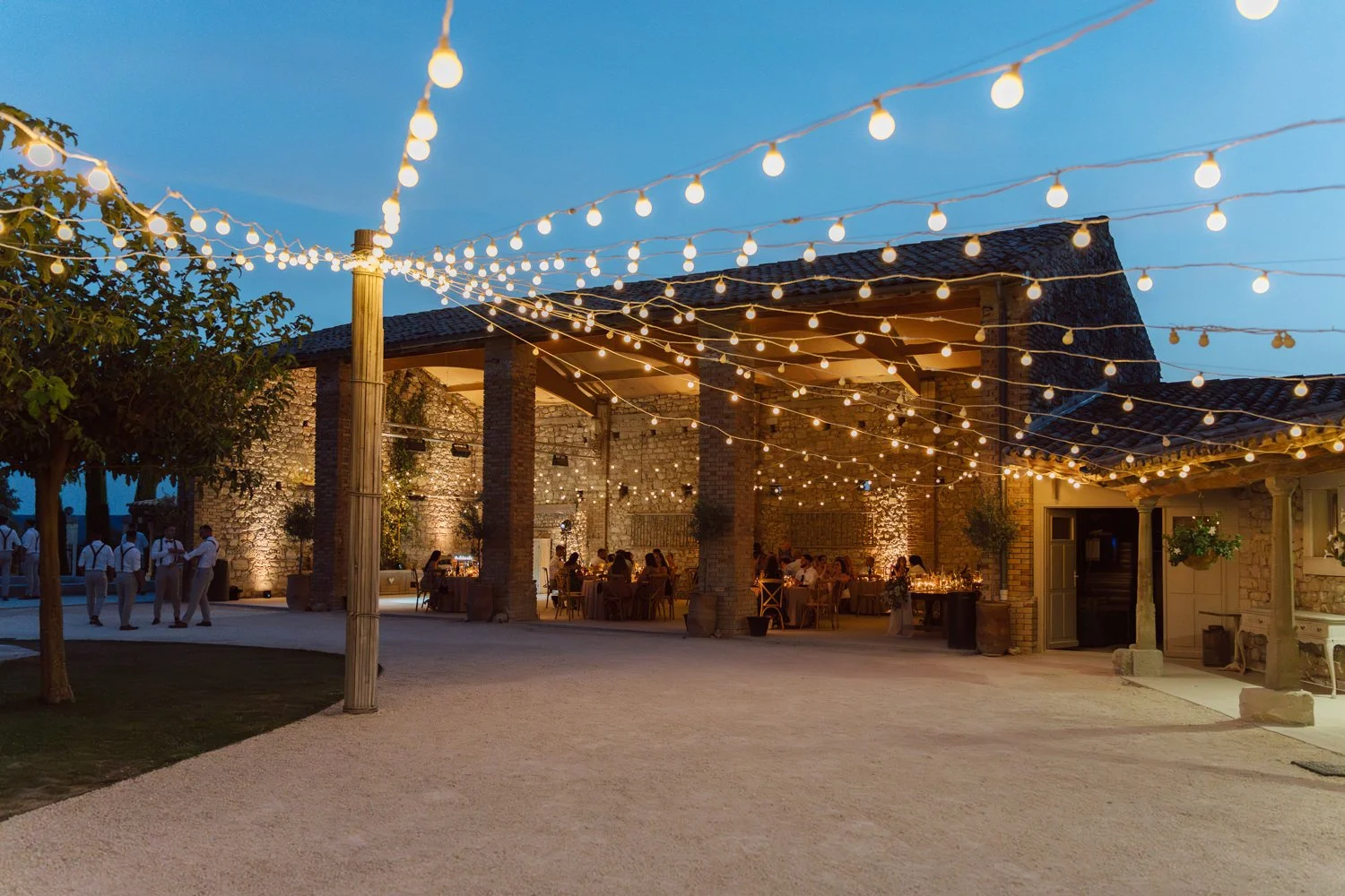Réception de mariage en soirée dans une cour en pierre illuminée par des guirlandes lumineuses, invités attablés sous une halle en Provence.