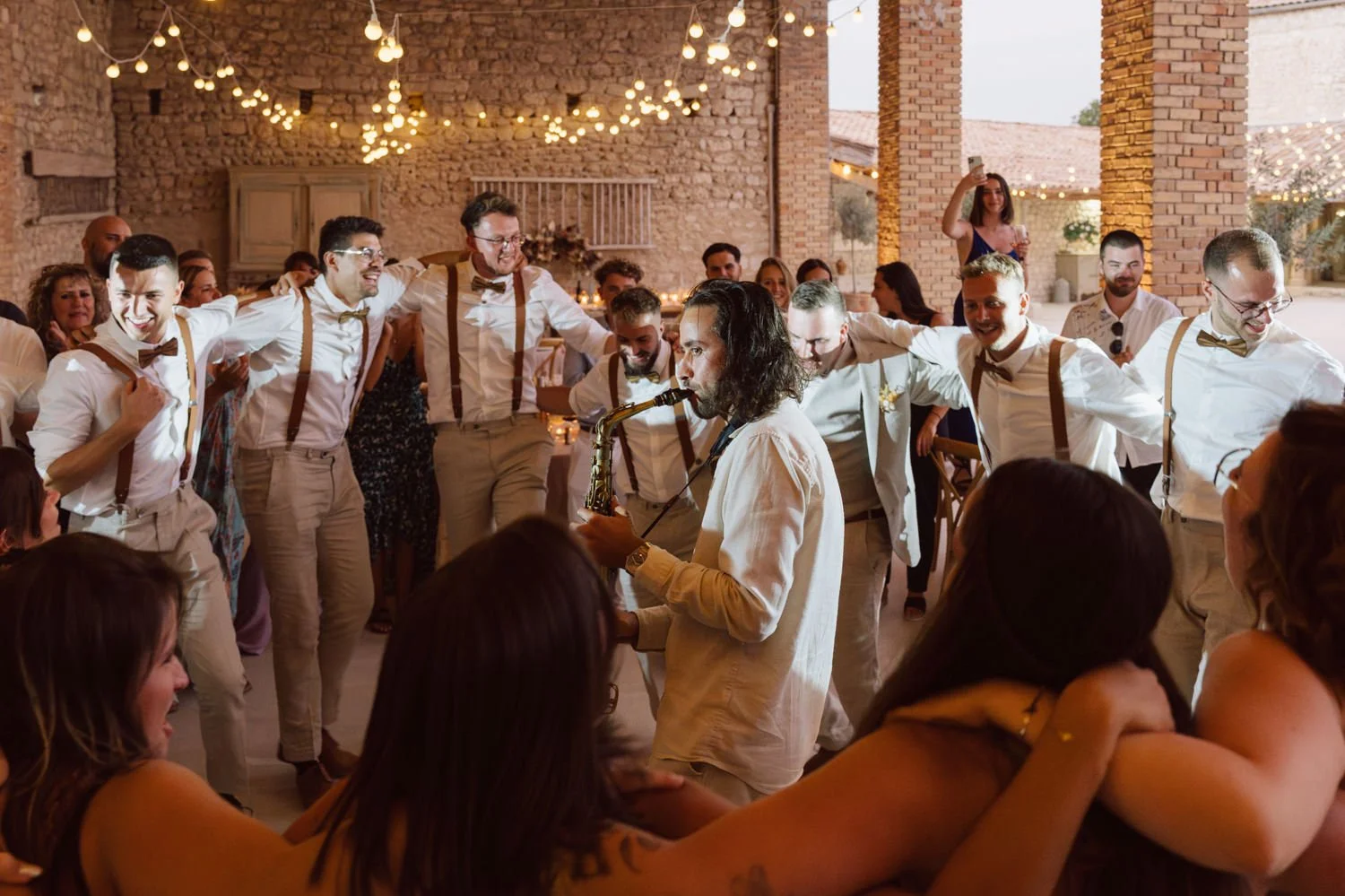 nvités et témoins entourent un saxophoniste sur la piste de danse lors de la soirée de mariage au Domaine de Sarson, sous des guirlandes lumineuses et des murs en pierre.