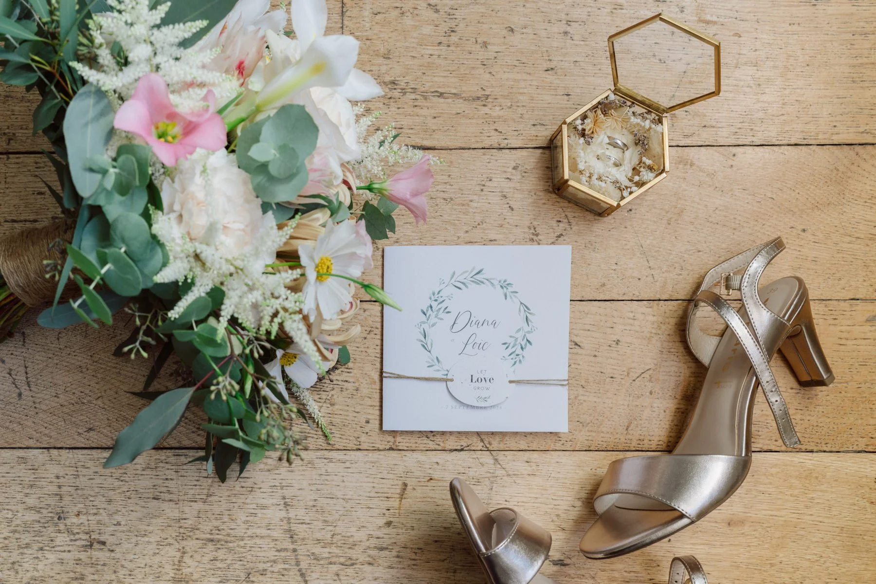 Flatlay des détails de mariage avec bouquet, papeterie, alliances et chaussures de la mariée, posé sur un sol en bois