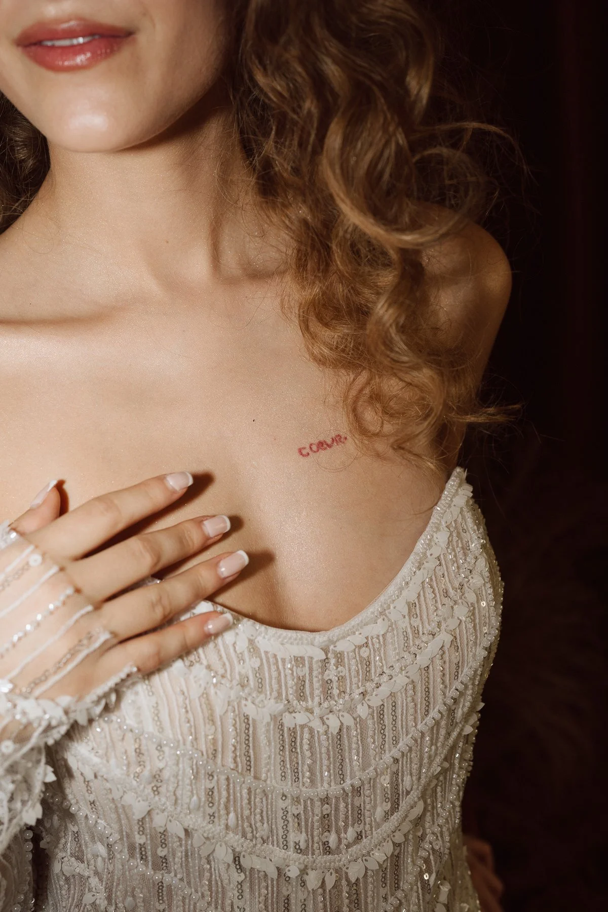 Détail de la robe de mariée perlée avec main posée sur le buste et petit tatouage cœur rouge sur la clavicule, photo de mariage au style éditorial lumineux