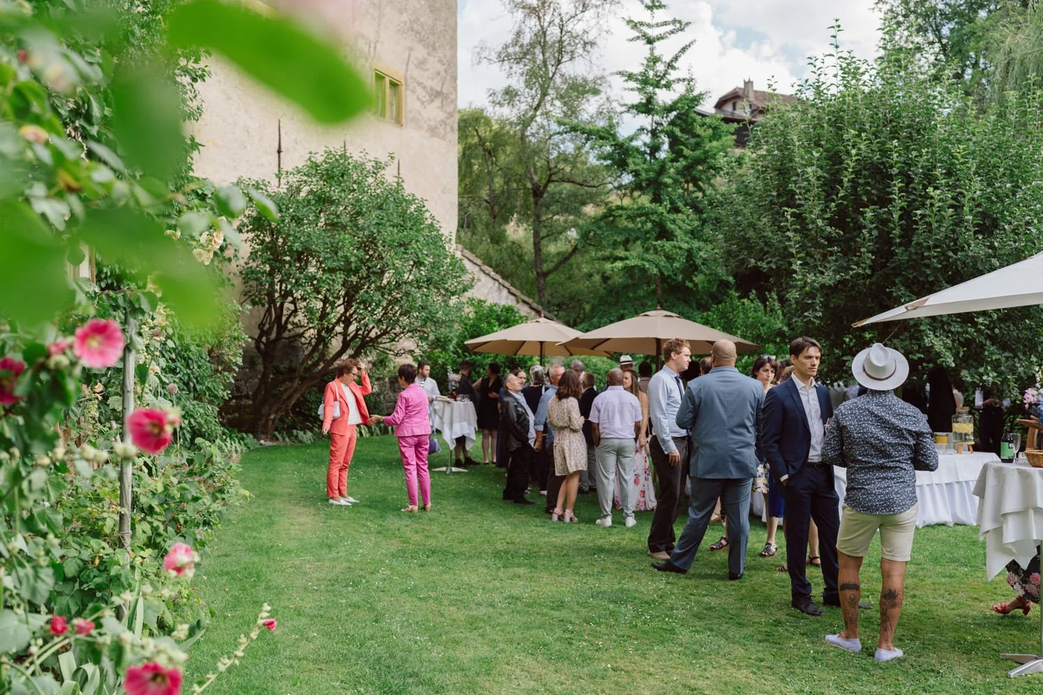 Gens d'affaires à une réception en plein air avec des parasols, dans un jardin verdoyant avec des arbres et des fleurs, devant un bâtiment ancien.