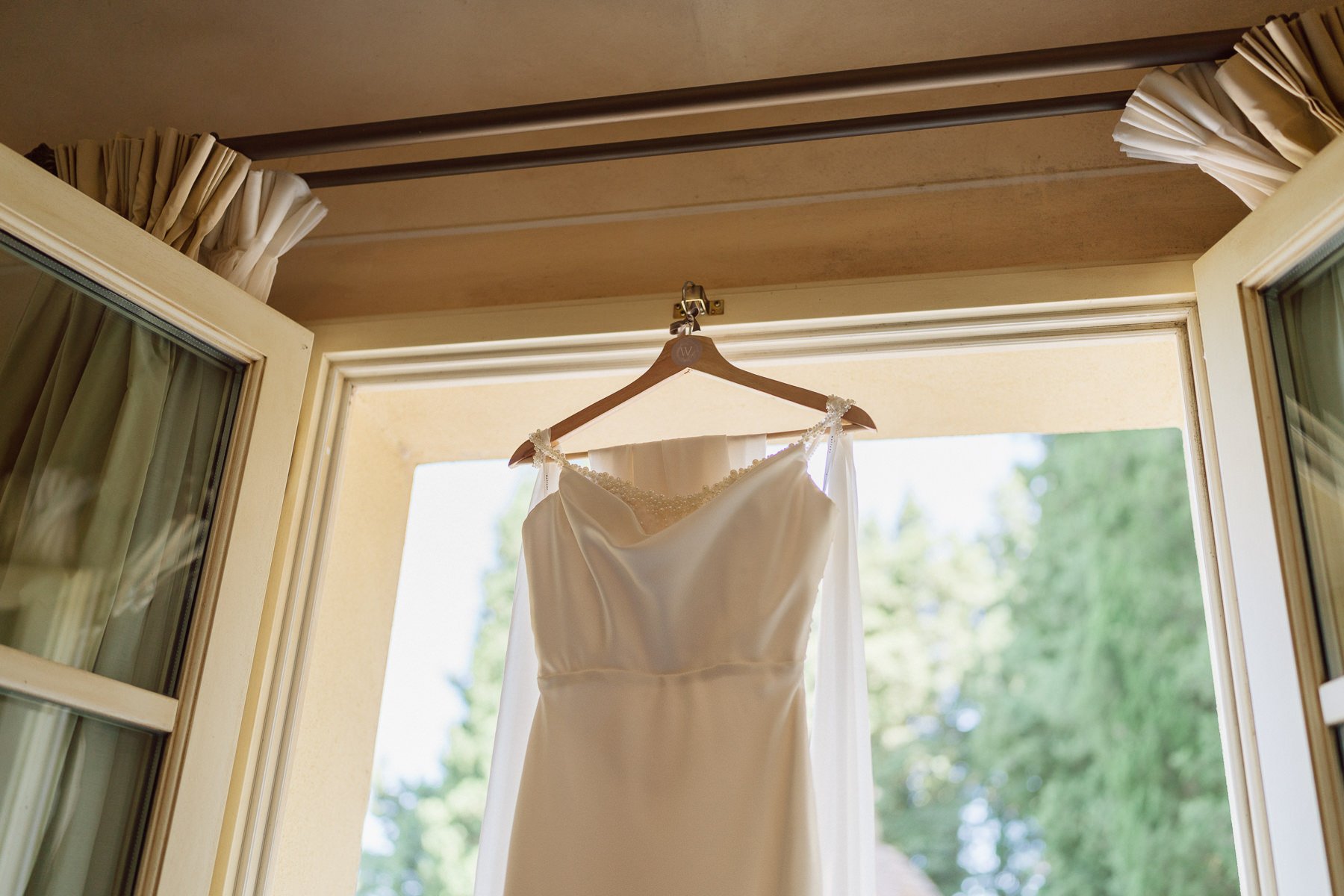 Robe de mariée suspendue devant une grande fenêtre baignée de lumière naturelle avec vue sur la verdure, détail poétique des préparatifs d’un mariage en Toscane