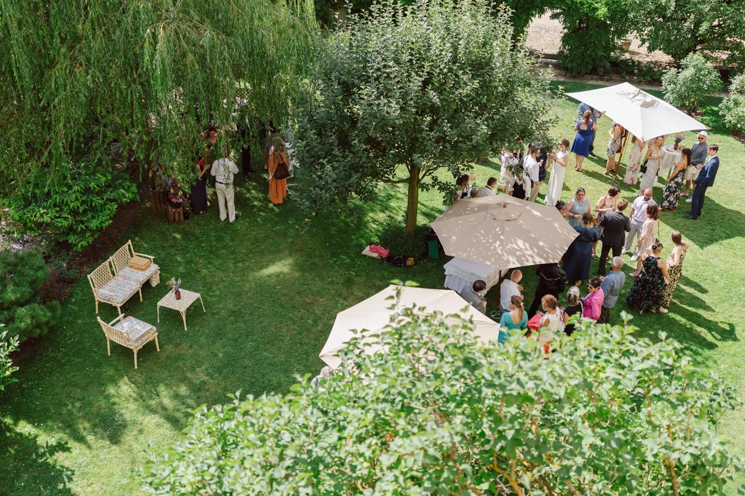 Vue aérienne du cocktail de mariage dans un jardin verdoyant, invités réunis sous des parasols à l’ombre des arbres, ambiance estivale et conviviale.