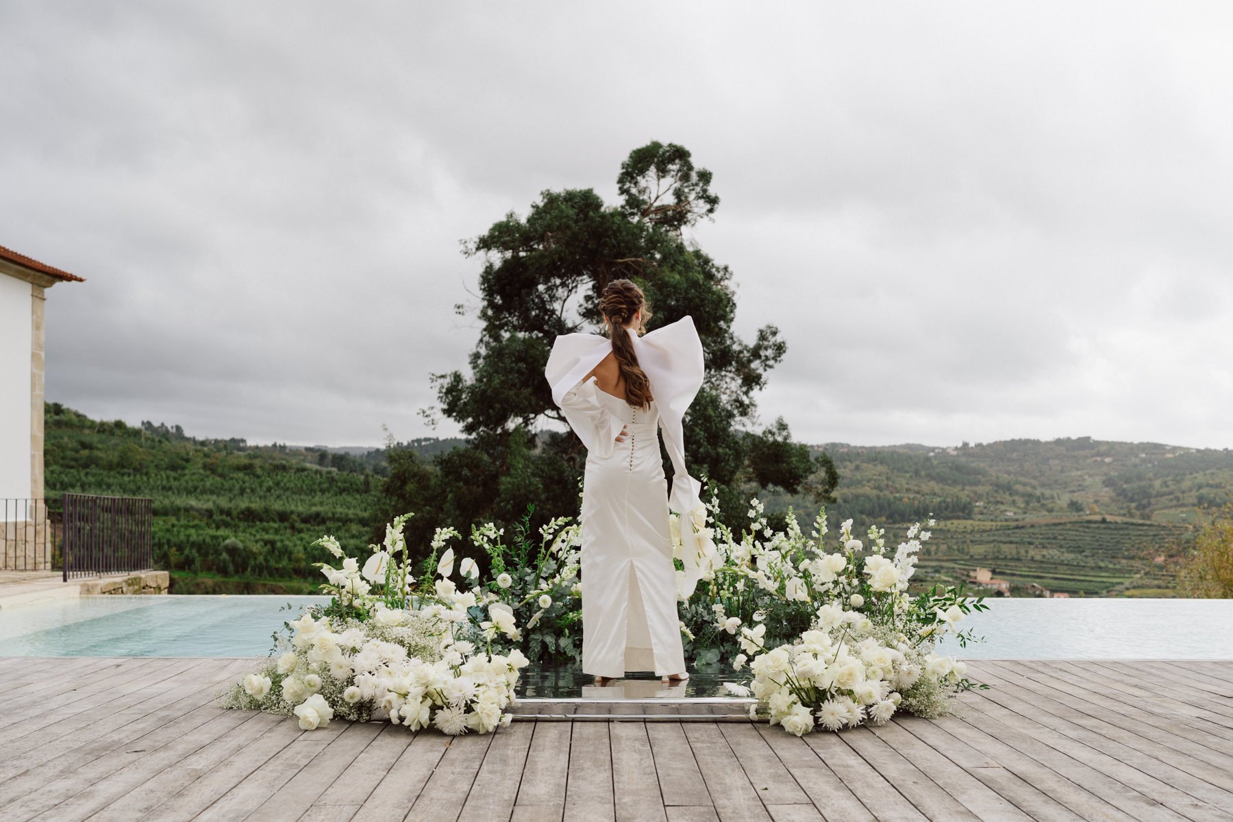 Mariée portant une robe Alma Real devant une arche florale blanche face aux collines de la Quinta do Pinho au Portugal – mariage destination élégant et raffiné