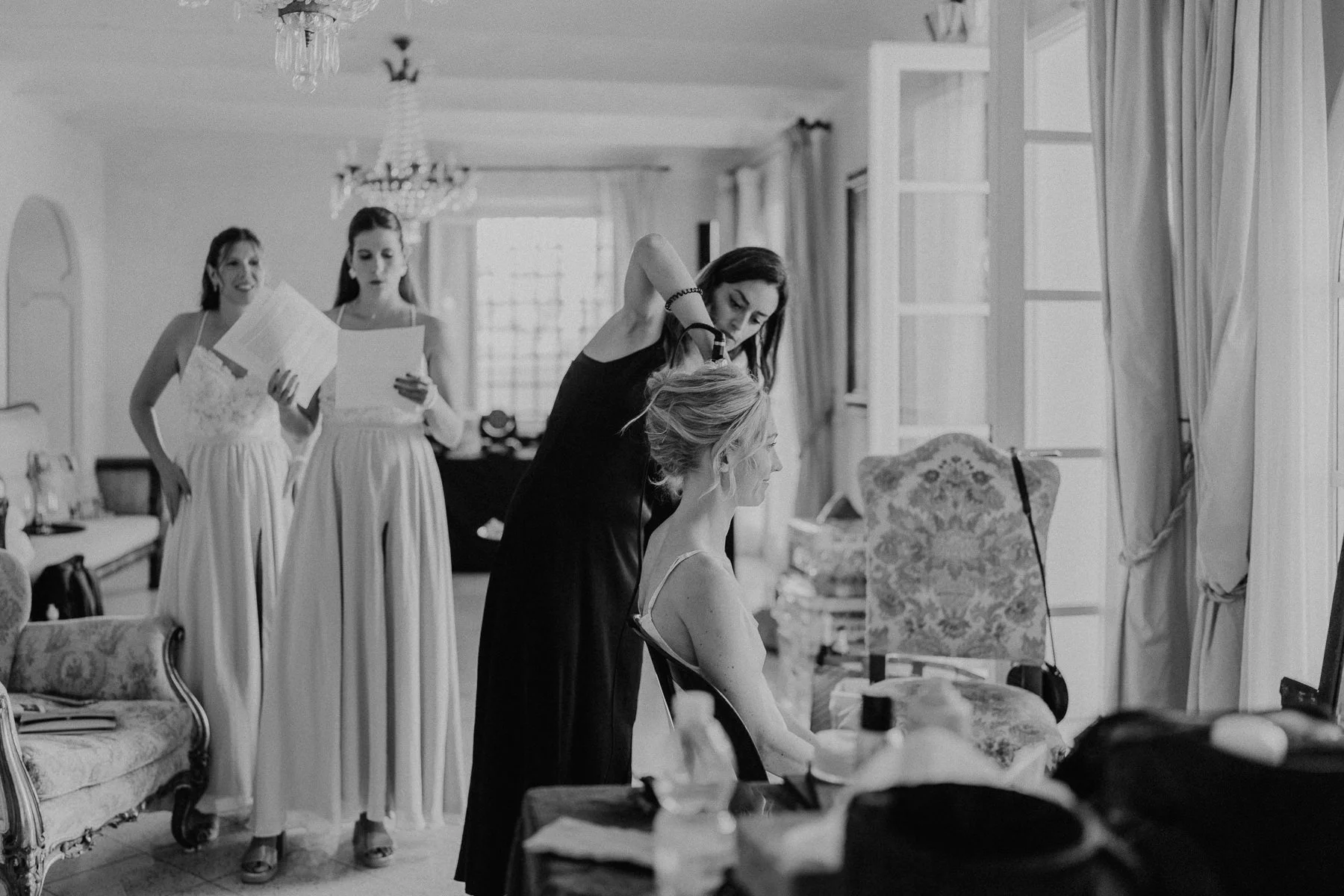 La mariée se fait coiffer pendant les préparatifs du mariage, tandis que ses demoiselles d’honneur lisent des discours dans une maison lumineuse en Toscane