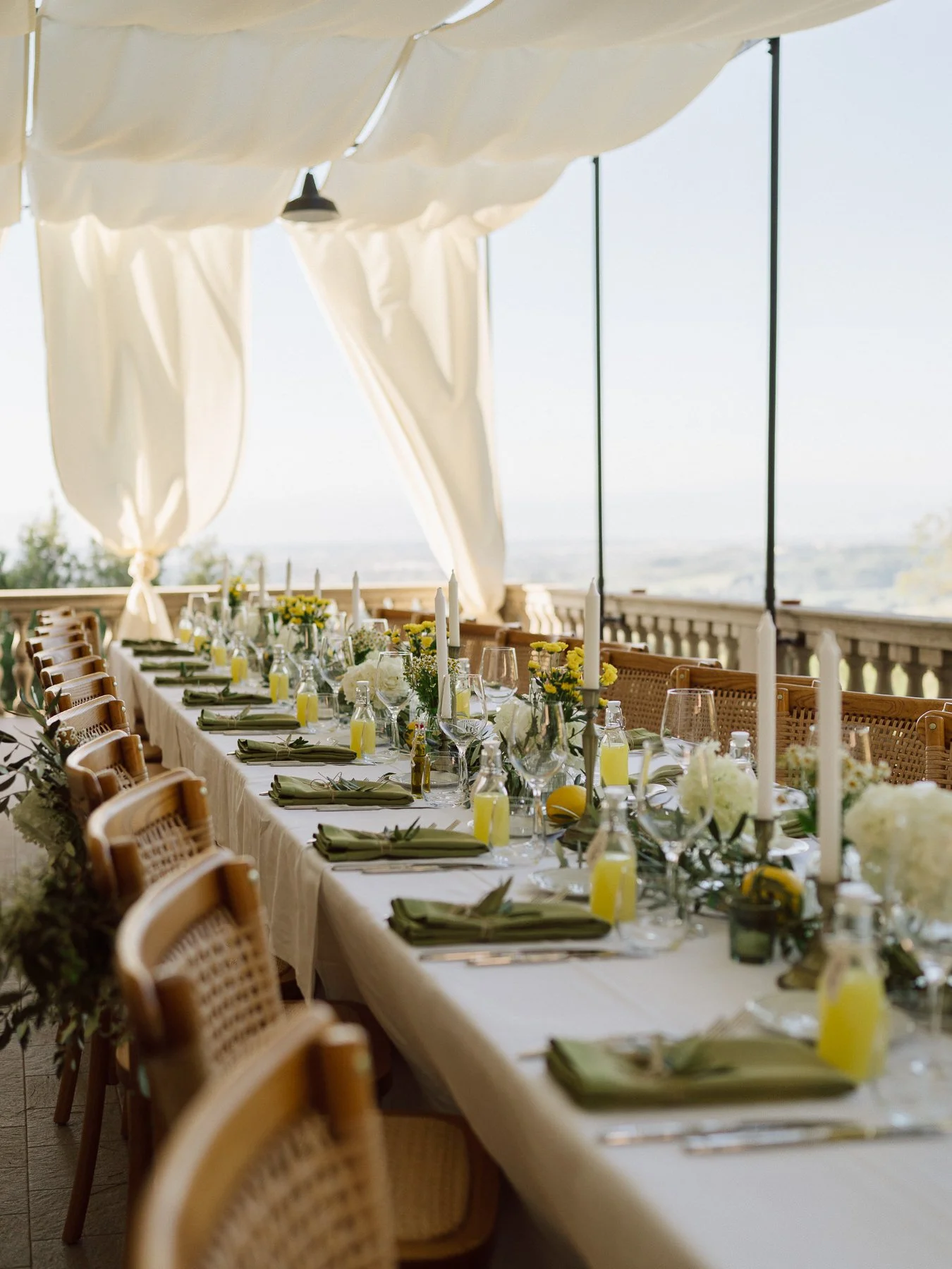 Grande table de réception de mariage décorée avec fleurs, bougies et serviettes vert olive sous une tente avec vue sur les collines toscanes