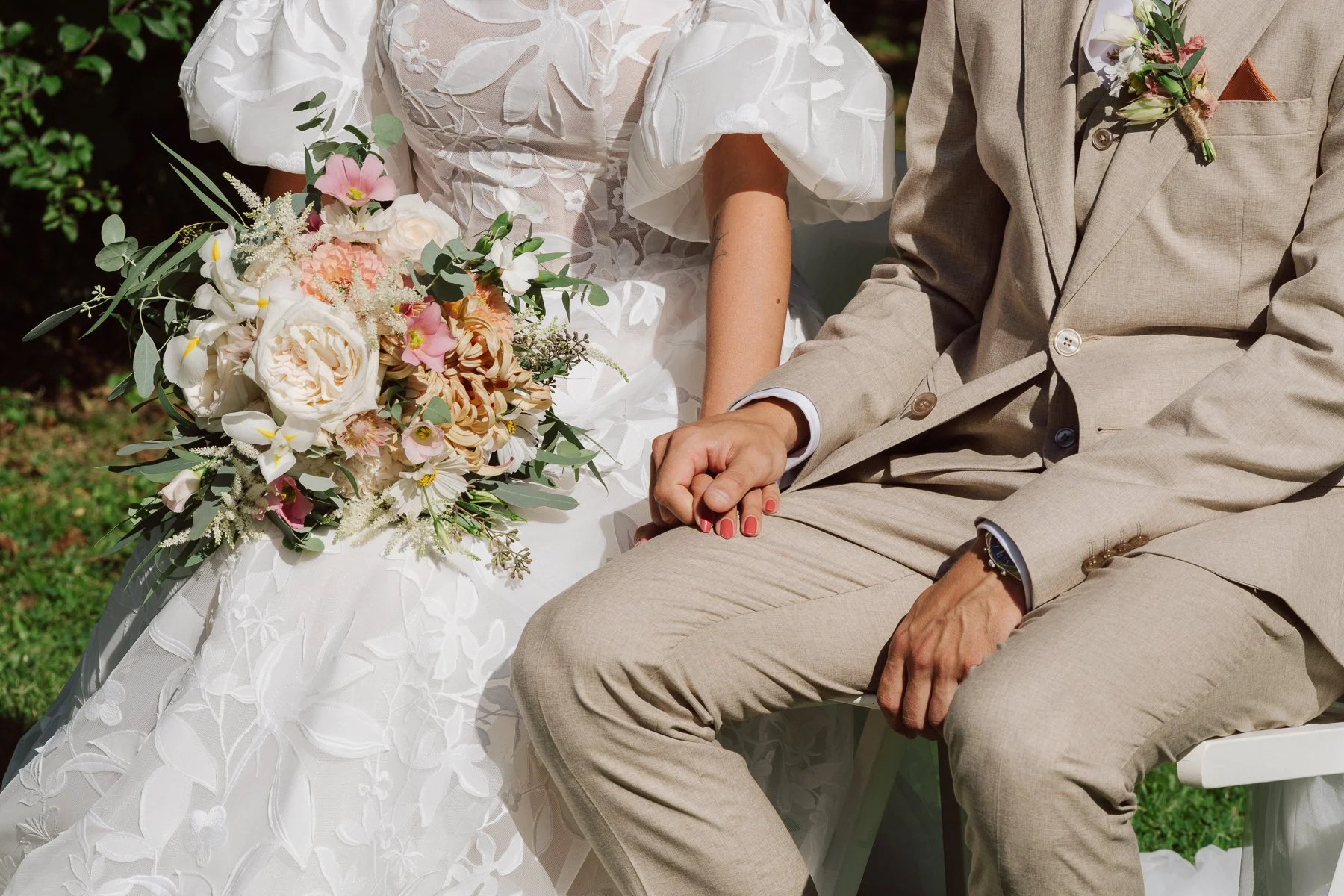 Détail des mariés assis côte à côte, mains qui se frôlent et bouquet de la mariée, lors du mariage de Diana et Loïc au Castel Bois-Genoud.