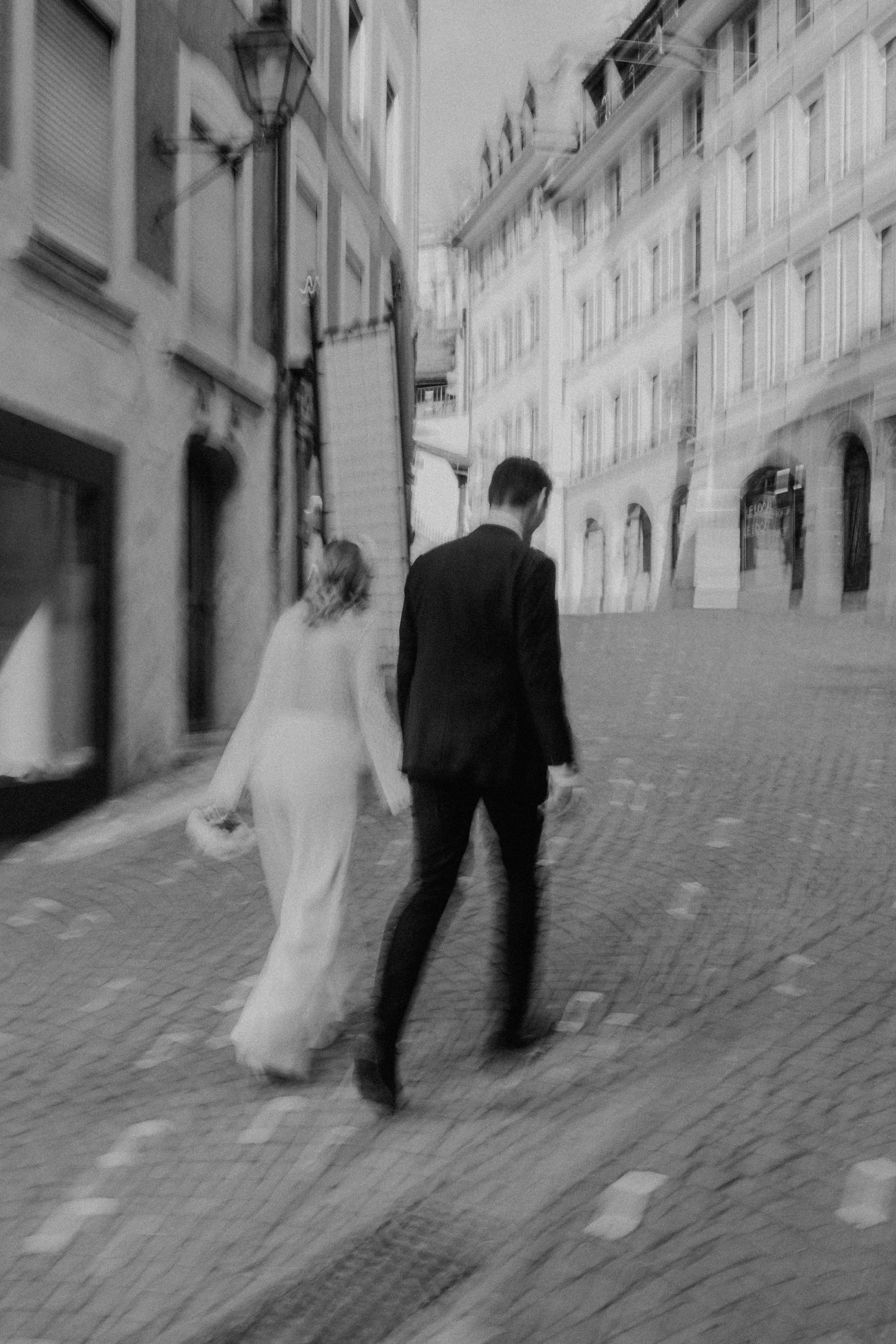 Un couple de mariés marche main dans la main dans une rue pavée au charme européen, entourée de bâtiments historiques. Le flou artistique de l’image ajoute une touche onirique et intemporelle, capturant l’essence d’un moment spontané et romantique da