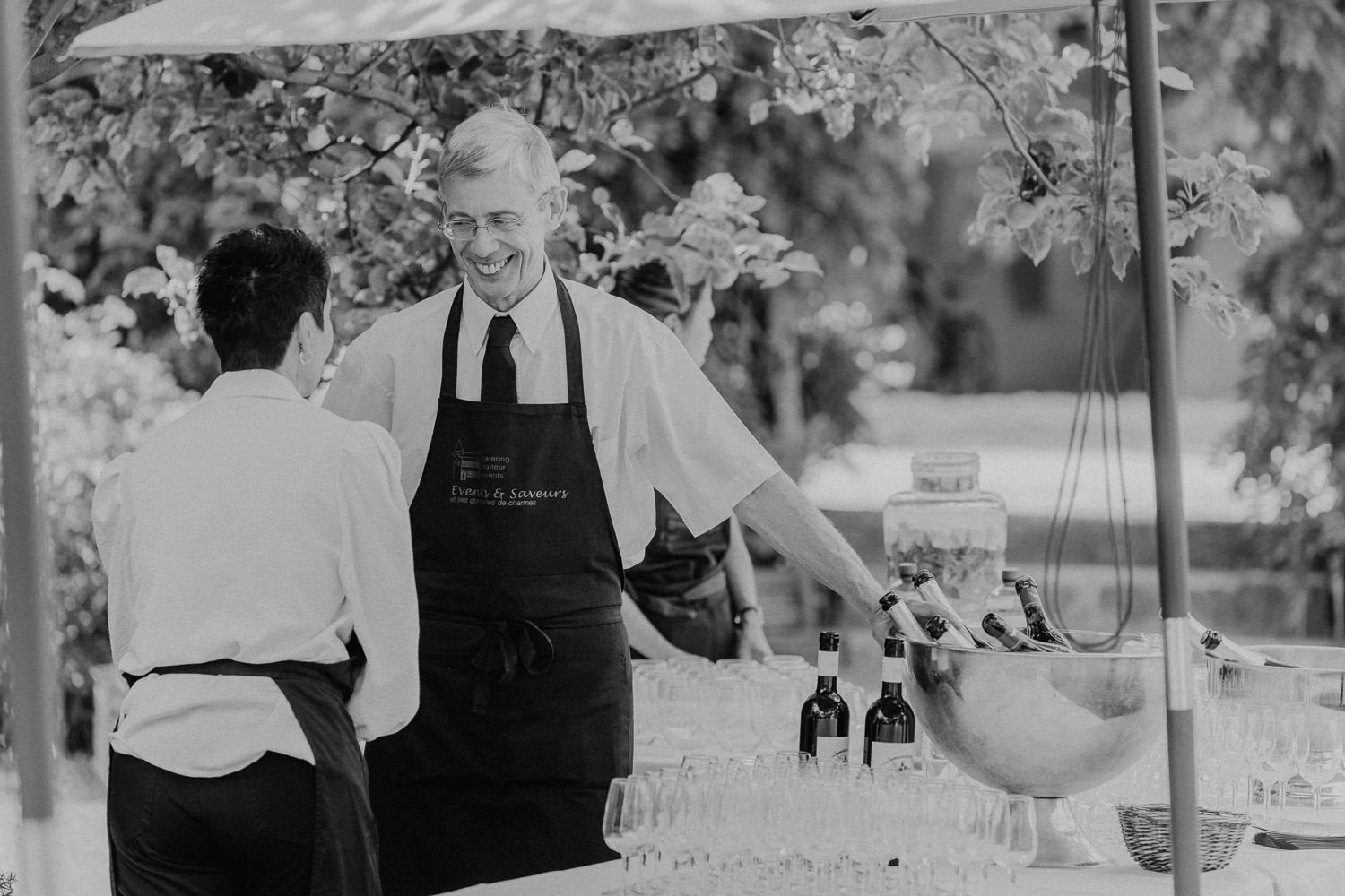 Service du cocktail lors d’un mariage au Jardin Baillival à Romainmôtier, équipe traiteur souriante derrière le bar, verres et bouteilles prêts à être servis, photo en noir et blanc par Nonante Émotions.