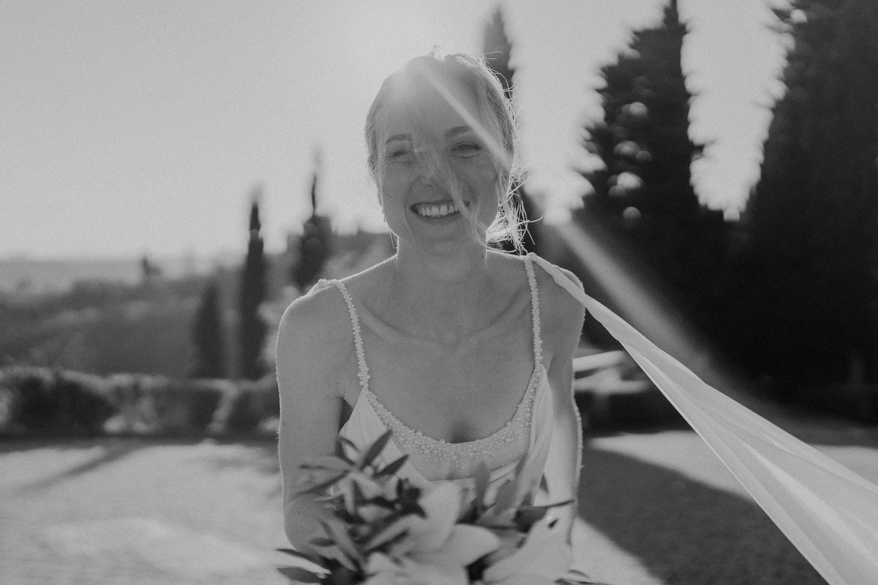 Une femme souriante portant une robe légère, en plein air, avec des arbres en arrière-plan, en noir et blanc.