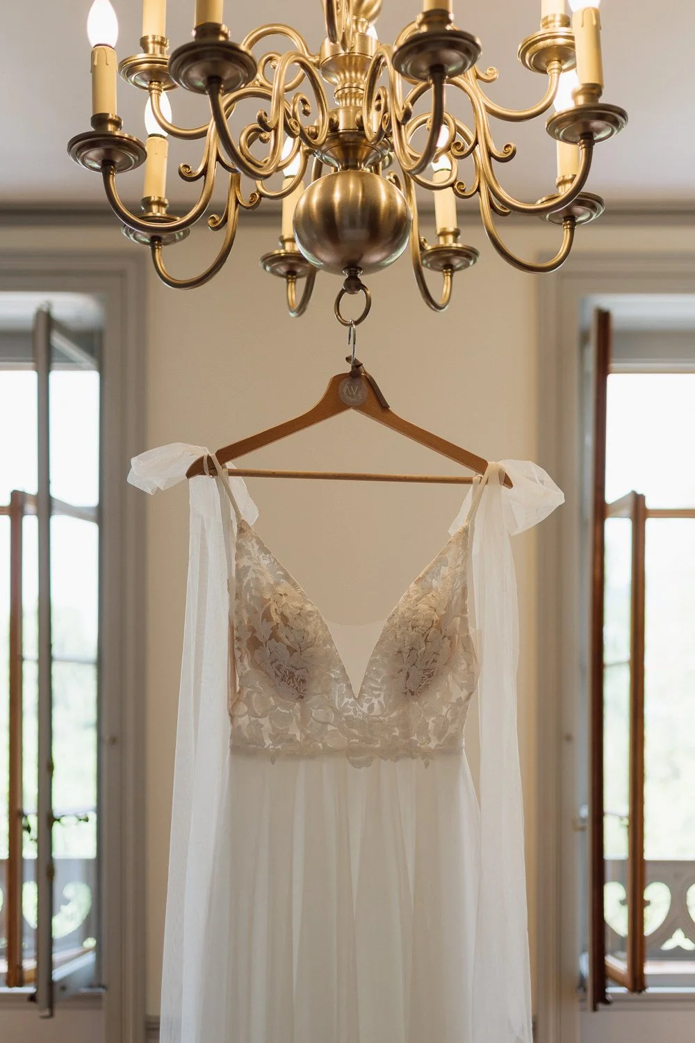 Robe de mariée signée With Love Boutique suspendue sous un lustre lors des préparatifs du mariage de Noémie et Bruno au Château de l’Aile à Vevey, photo de mariage par Nonante Émotions