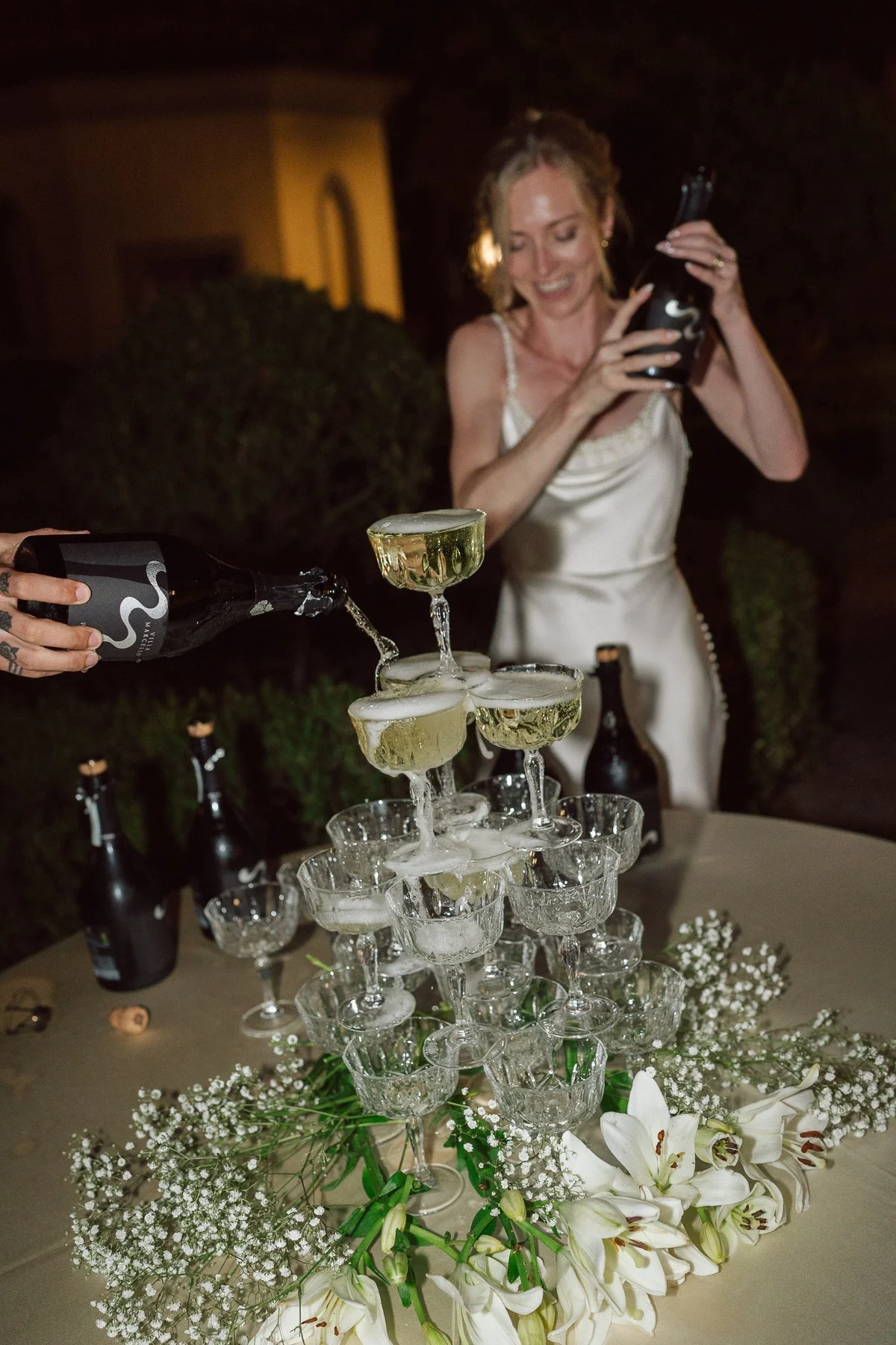 Mariée souriante versant du champagne sur une pyramide de coupes lors de la soirée de mariage, décor floral blanc et ambiance festive de nuit en Toscane.