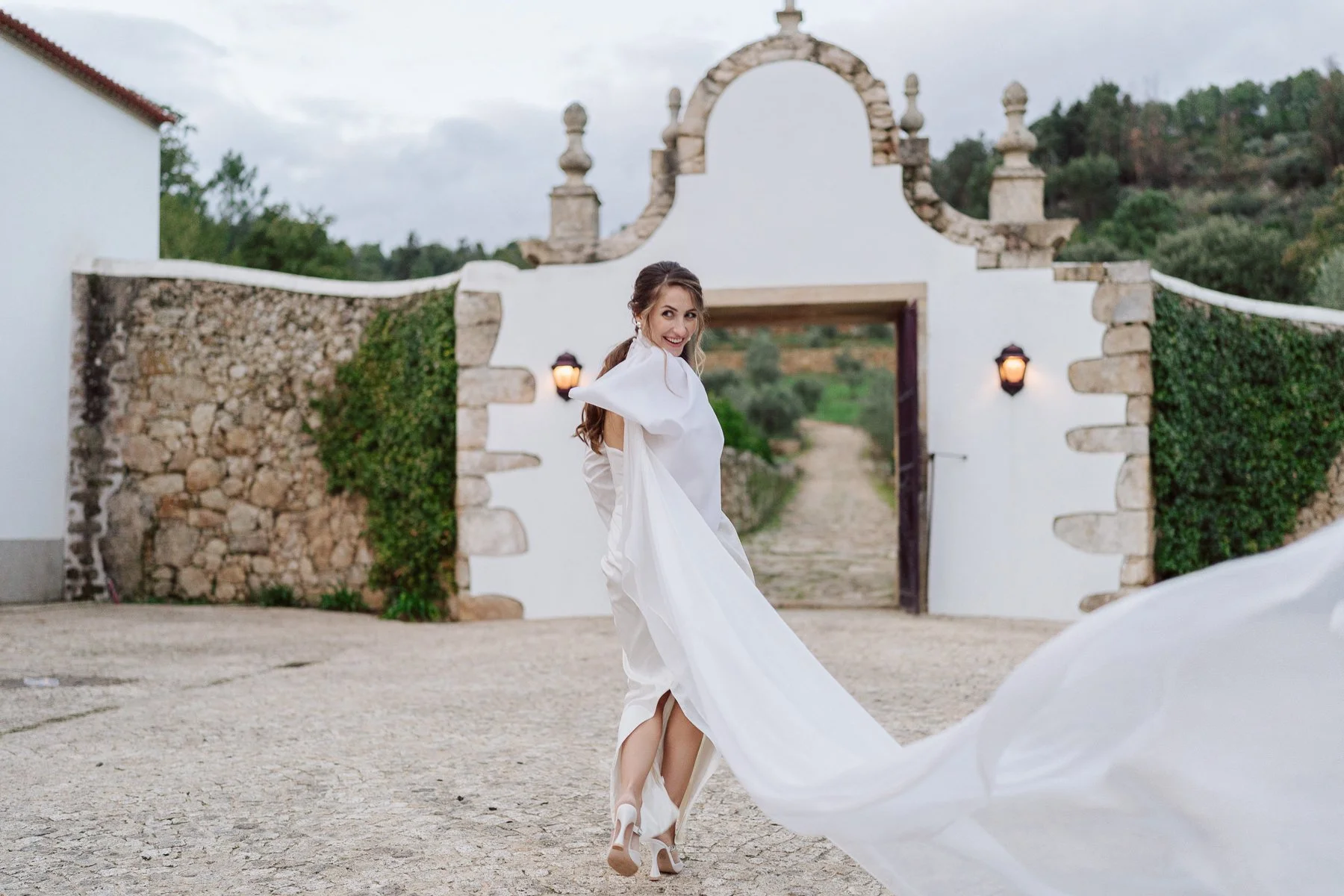 Mariée en robe fluide avec traîne au vent devant l’entrée d’un domaine en pierre, photo de couple élégante et naturelle dans un cadre méditerranéen