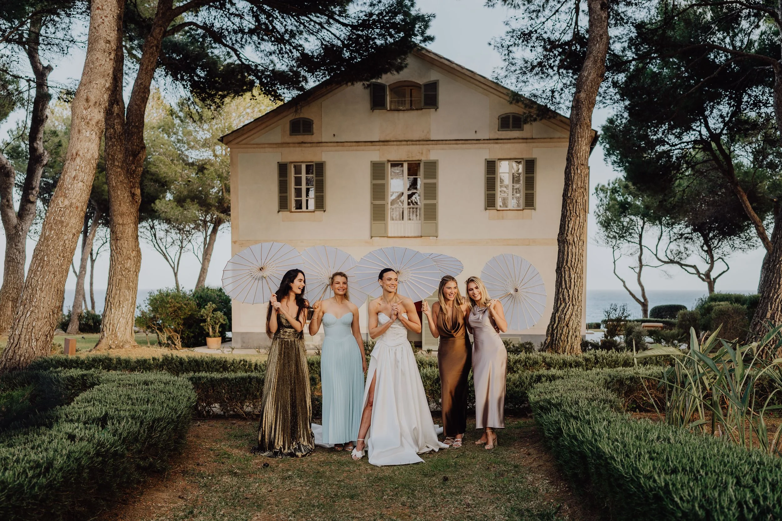 Instant de complicité entre la mariée et ses demoiselles d’honneur, réunies sous des ombrelles dans un jardin baigné de lumière d’été.