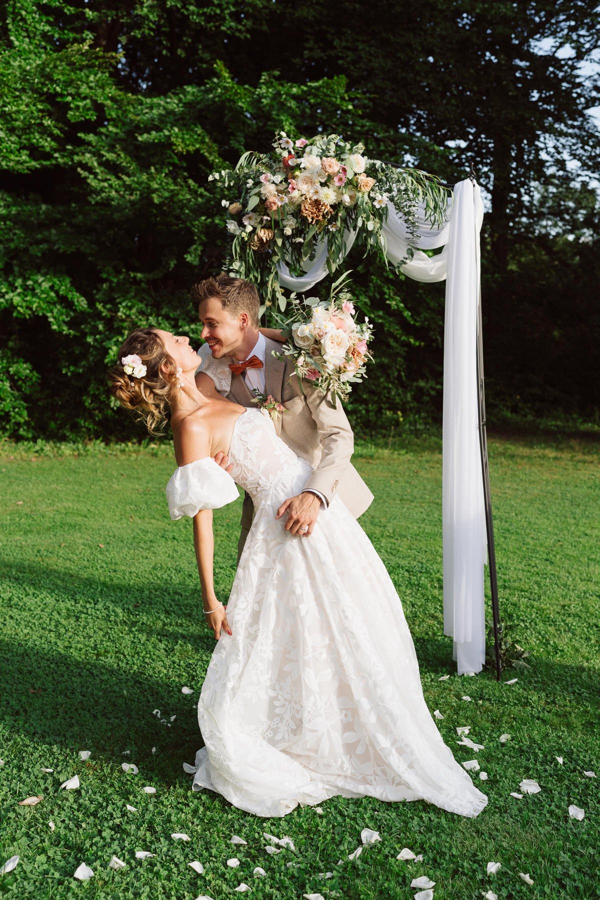 Couple de mariés s’embrassant sous une arche fleurie lors d’une cérémonie de mariage en plein air, décor floral élégant dans un jardin verdoyant, photo naturelle et émotionnelle par Nonante Émotions