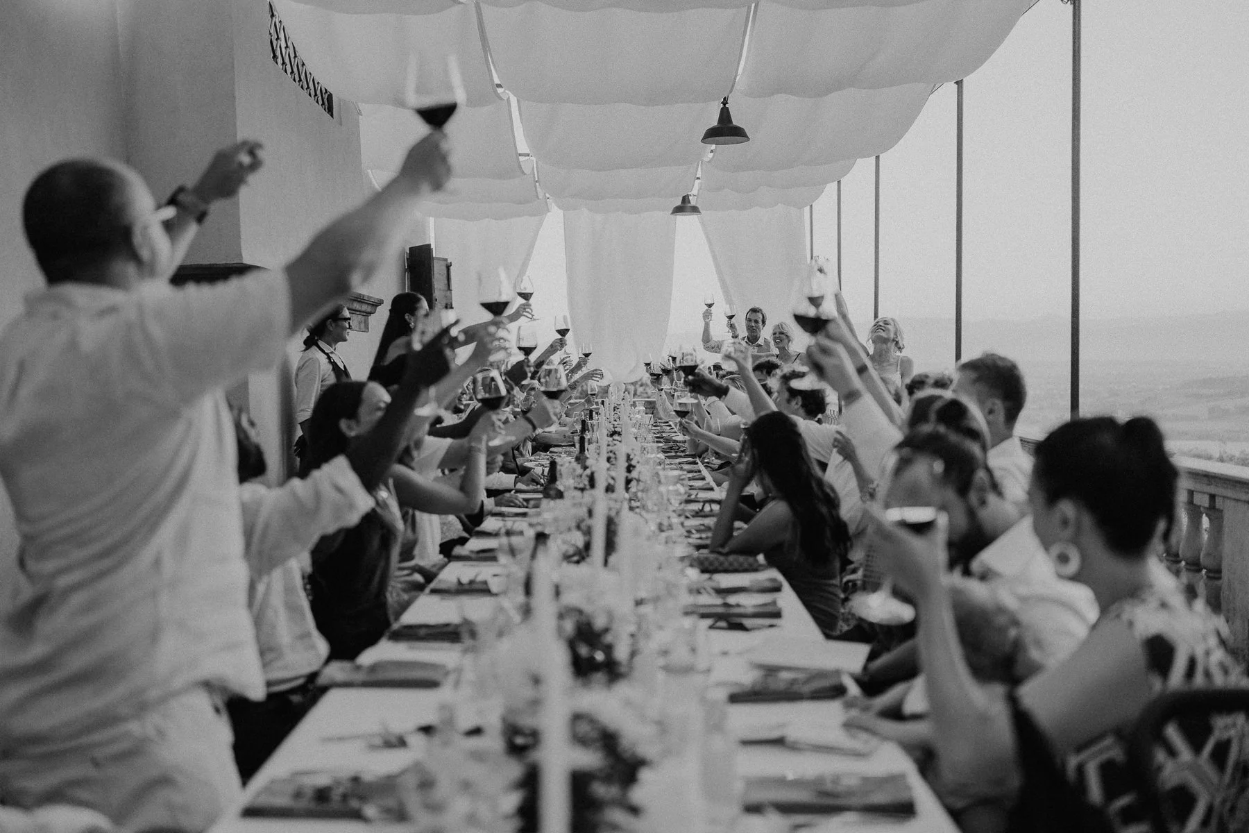 nvités levant leurs verres lors d’un toast pendant un dîner de mariage sur une longue table en terrasse en Toscane, photo en noir et blanc avec vue sur les collines.