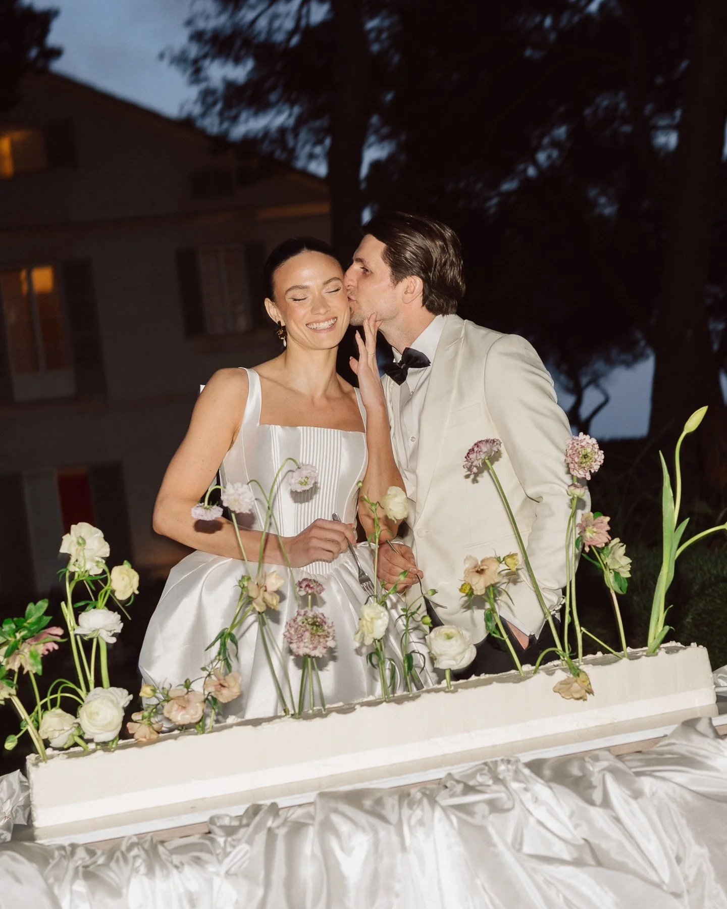 Bride and groom cutting their wedding cake during an elegant evening reception in Mallorca, surrounded by delicate floral arrangements and a modern luxury wedding atmosphere