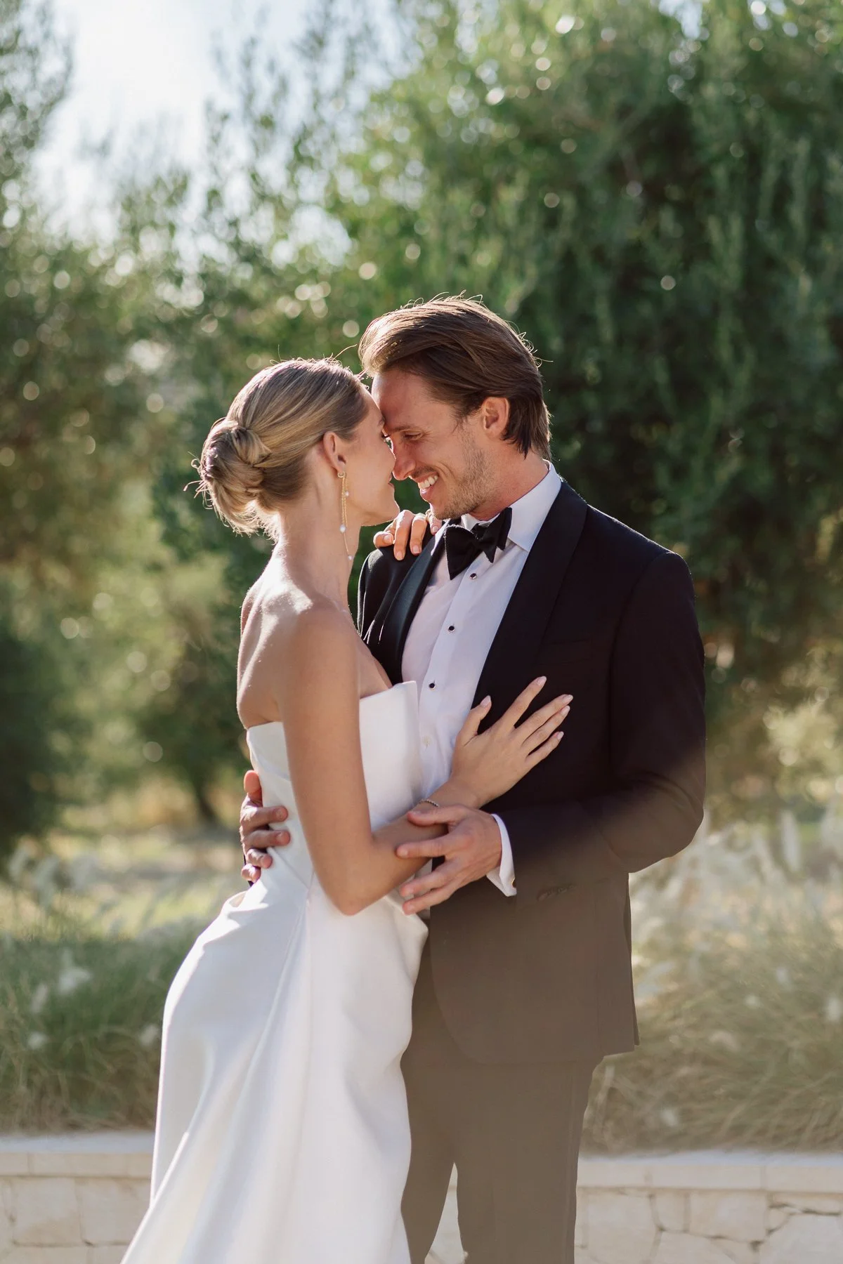 Un couple de mariés souriant en se tenant dans un cadre extérieur avec des arbres en arrière-plan.
