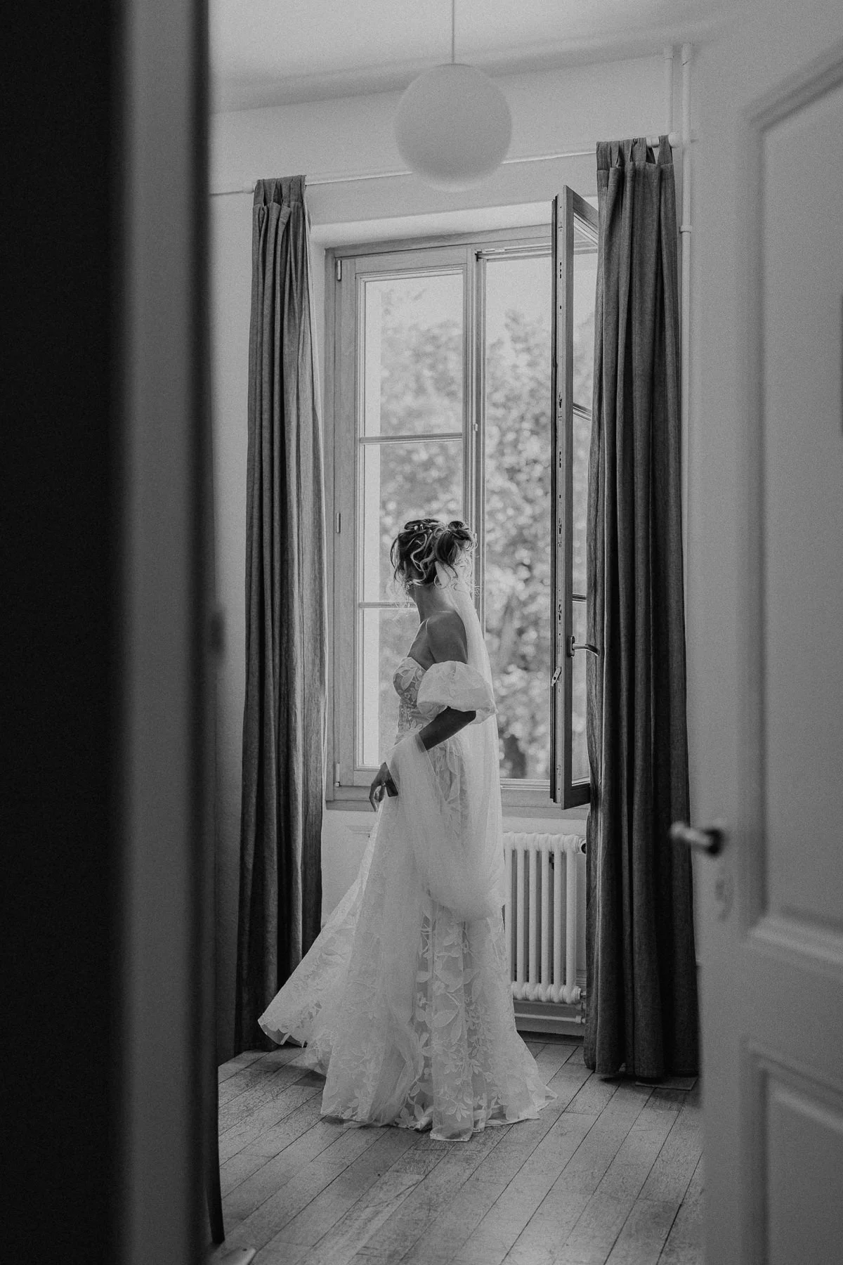Mariée en robe de mariage se tenant près d’une fenêtre pendant ses préparatifs au Castel Bois-Genoud.