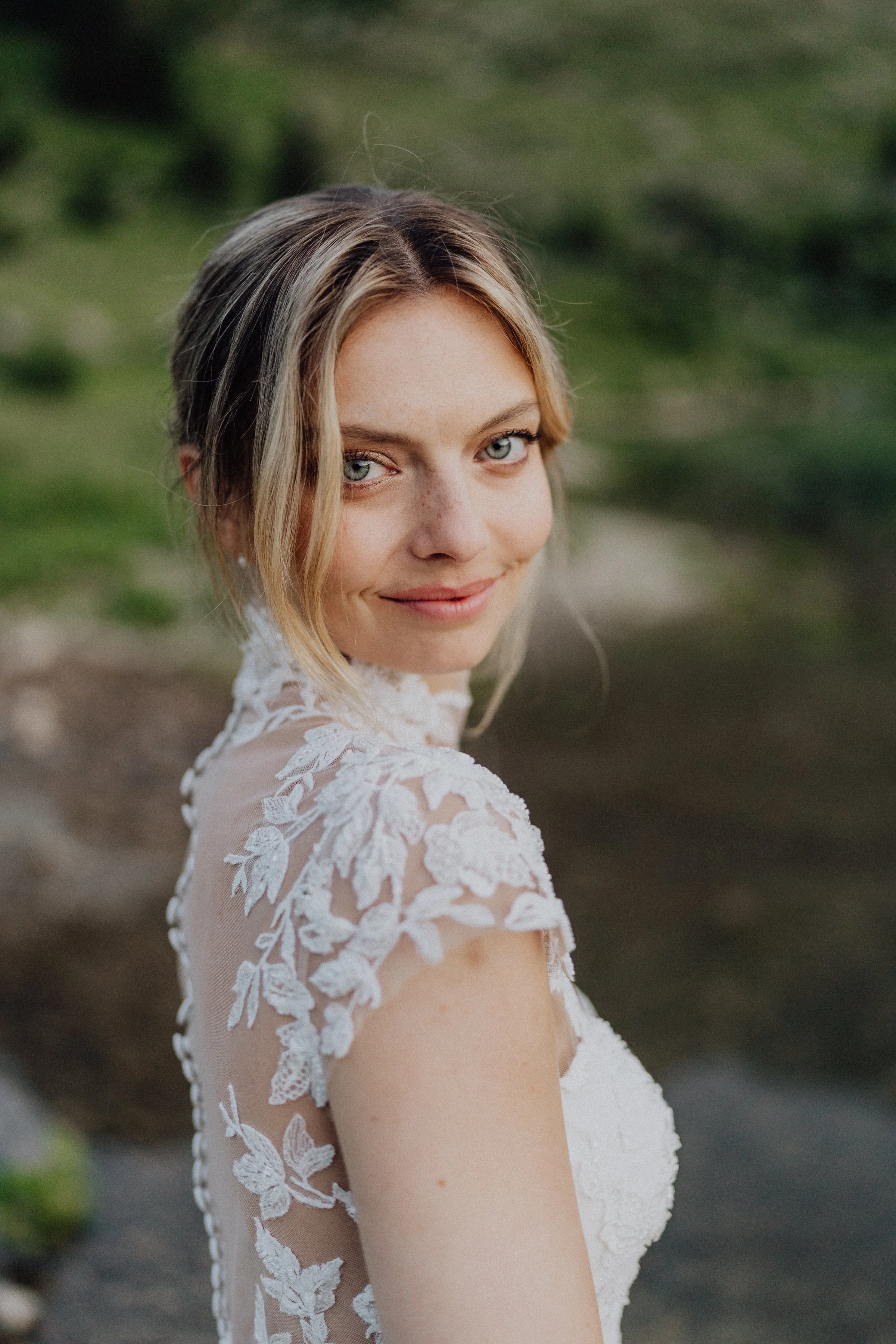 Un portrait naturel de la mariée avec un regard captivant. Sa robe en dentelle florale et les détails délicats ajoutent une touche romantique, tandis que l’arrière-plan verdoyant crée une ambiance apaisante et organique.