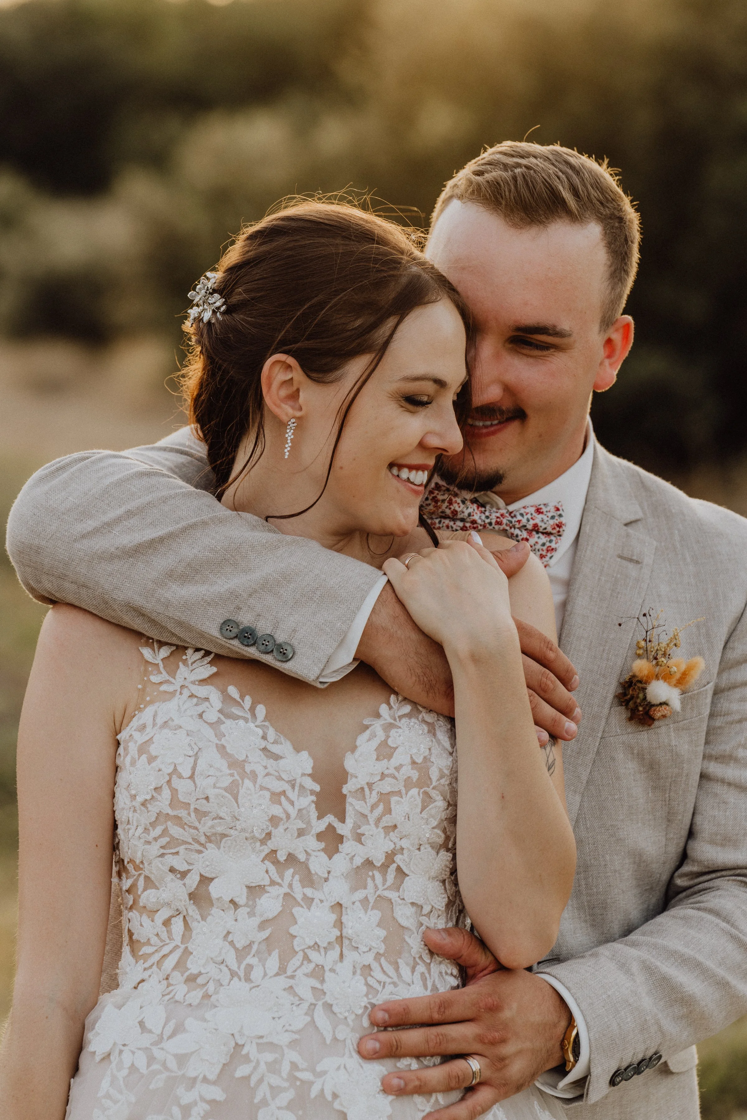  Un moment de tendresse entre les mariés, capturé à la lumière dorée du soleil au domaine de Sarson. La mariée porte une robe en dentelle délicate, et le marié, dans un costume en lin clair, enlace sa bien-aimée avec douceur. L'ambiance romantique et
