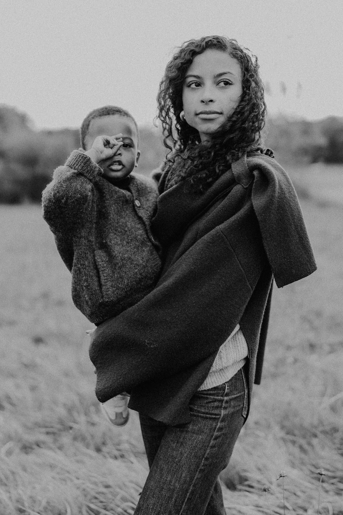 Une femme tient un garçon dans un champ naturel, tous deux regardant vers l'objectif, en noir et blanc.
