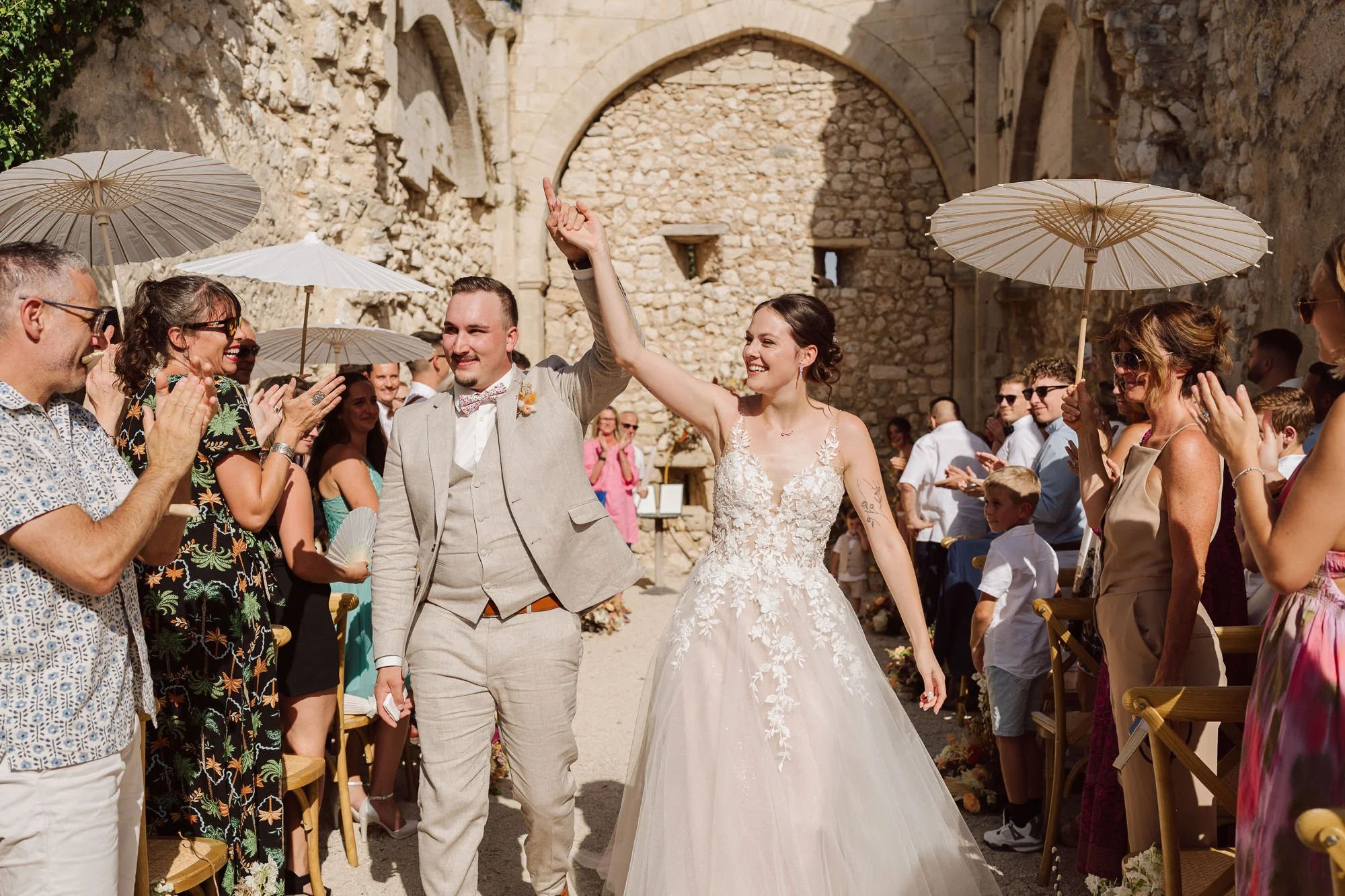 Cinthya et Alexis sortent de leur cérémonie laïque dans l’ancienne chapelle du Domaine de Sarson en Provence, sous les applaudissements de leurs invités tenant des ombrelles blanches.