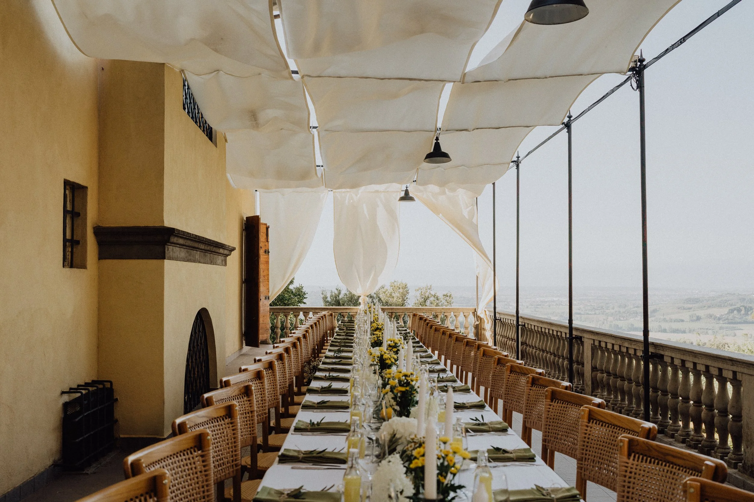 Une table de réception longue et élégante, dressée sur une terrasse avec vue panoramique sur la campagne toscane. Les nappes blanches, les décorations florales jaunes et blanches, et les chaises en osier tressé s’harmonisent sous une pergola ornée de