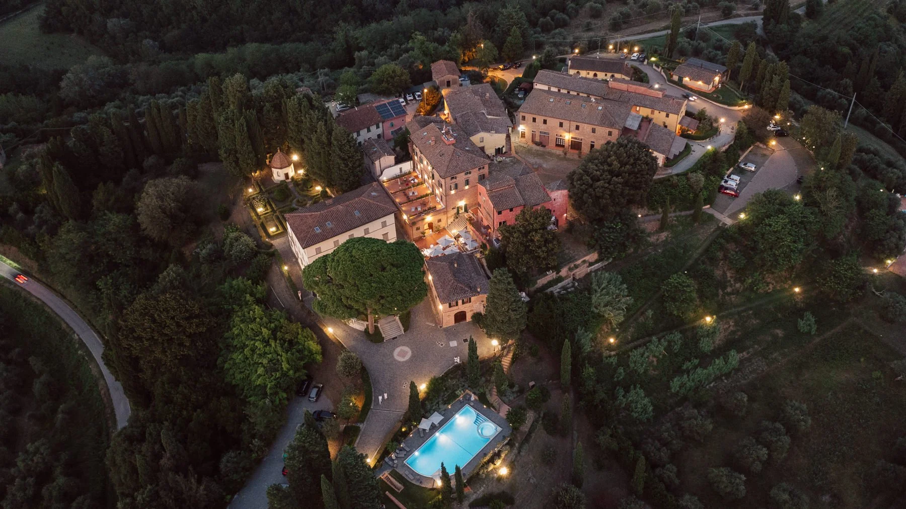 Vue drone de nuit du Borgo Bucciano en Toscane illuminé, lieu de mariage entouré de collines, cyprès et piscine éclairée.
