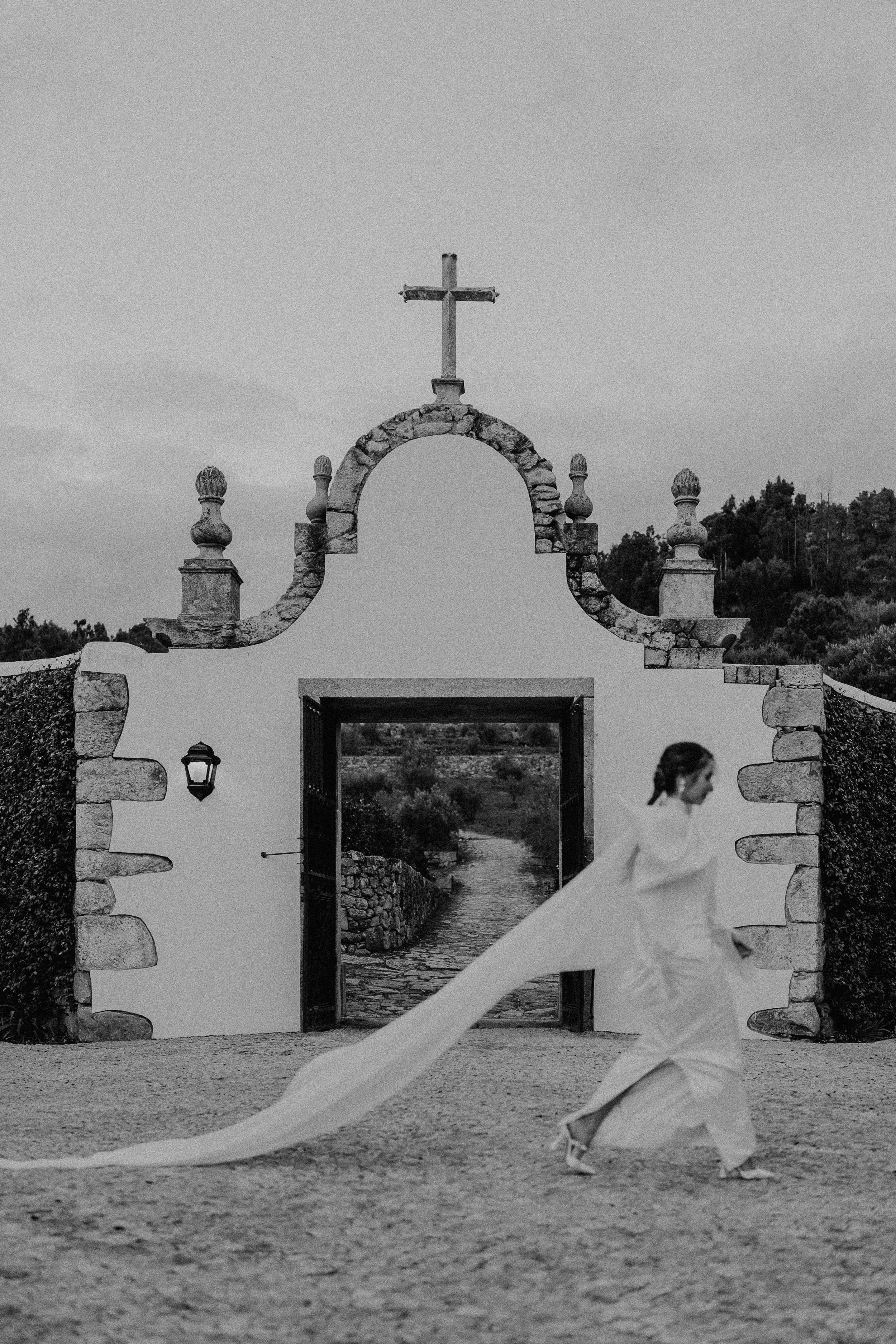 Une mariée élégante et moderne porte une robe Alma Real, mettant en valeur des lignes épurées et une traîne majestueuse qui flotte délicatement au vent. Devant la chapelle emblématique du domaine Quinta do Pinho, cette scène respire le raffinement et