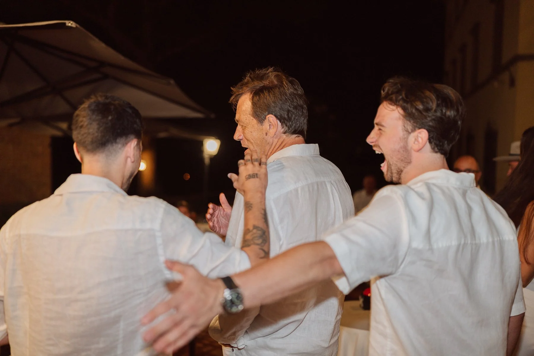 nvités masculins riant et marchant bras dessus bras dessous pendant la soirée d’un mariage en Italie, ambiance festive et chaleureuse.