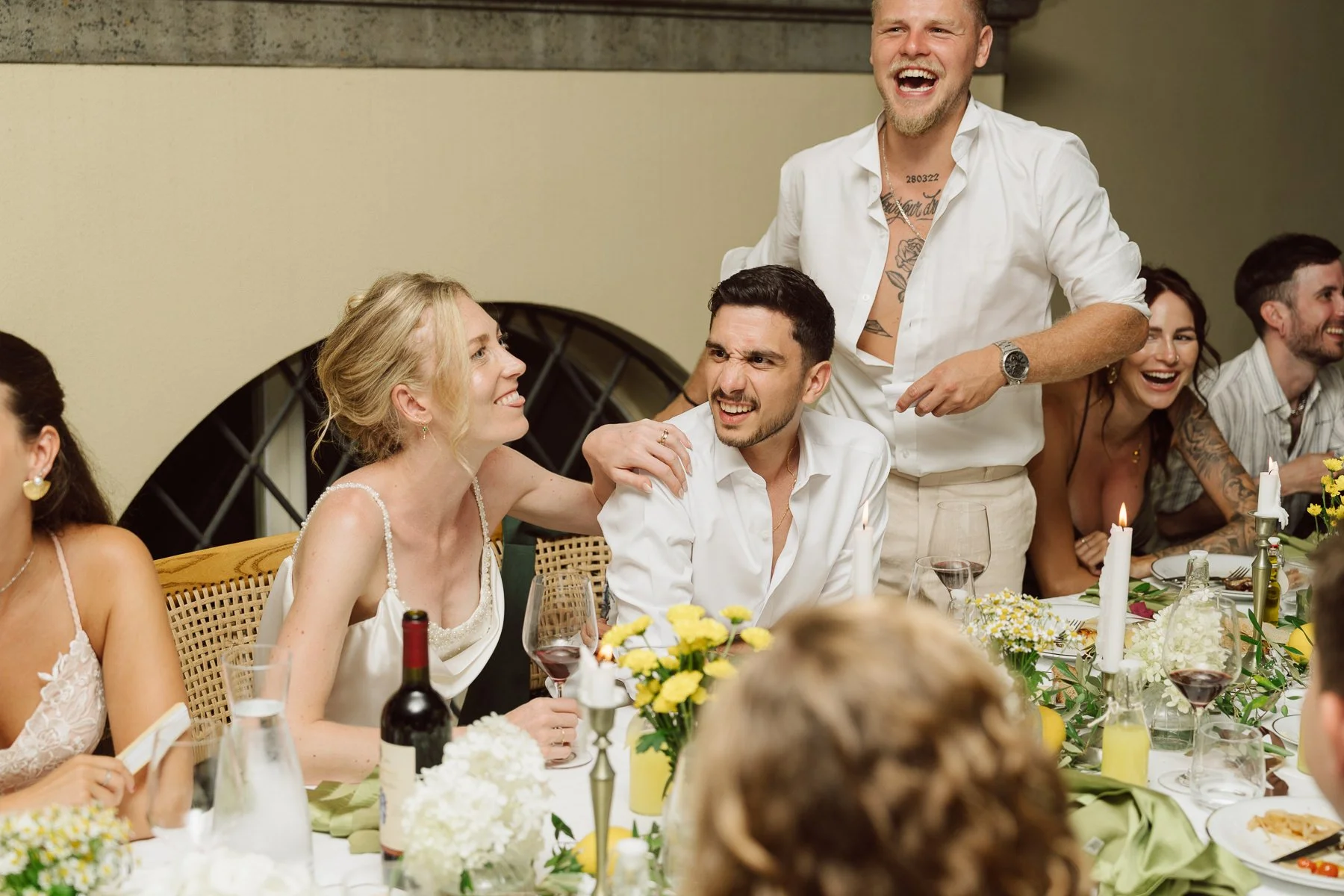 Mariés et invités riant pendant un discours de témoins lors du dîner de mariage, autour d’une table décorée de fleurs blanches, bougies et citrons, ambiance chaleureuse en Toscane.