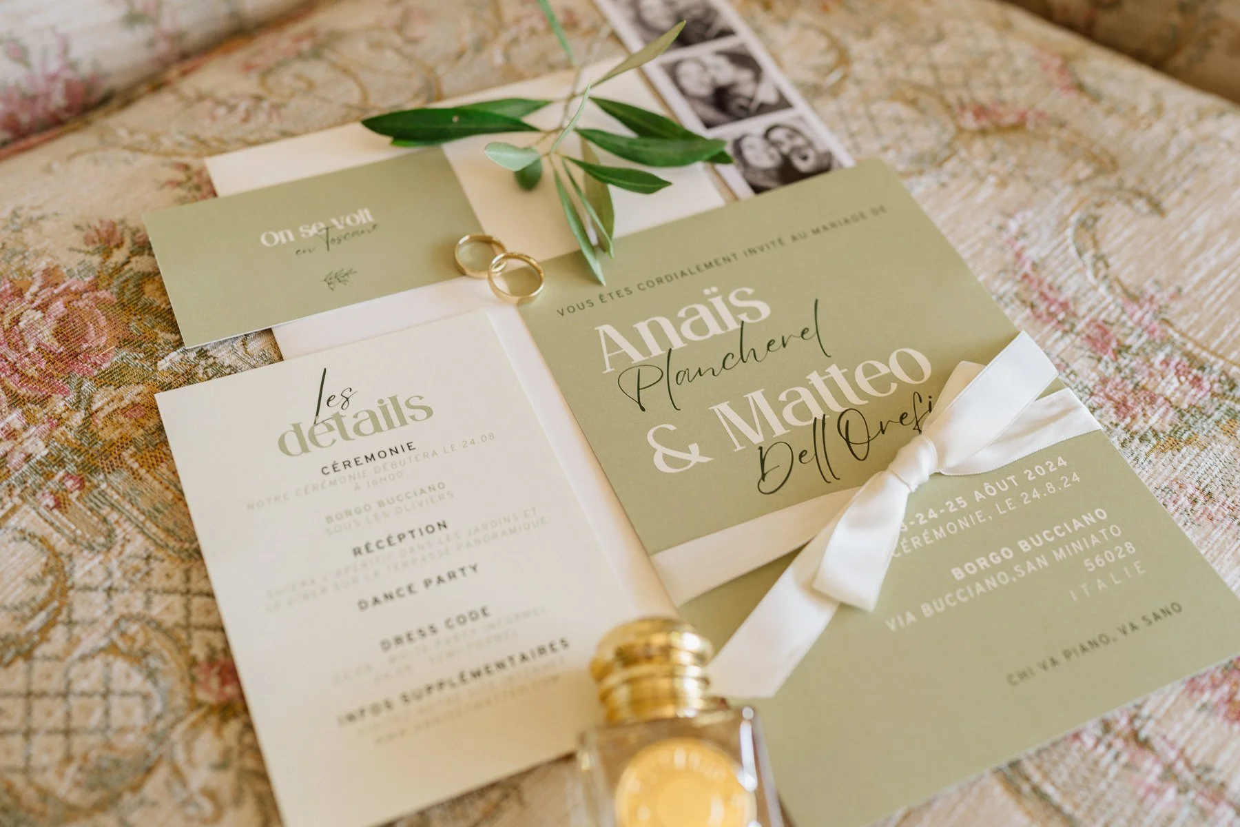 Faire-part de mariage vert olive avec alliances et ruban en soie, posé sur un tissu fleuri, pour un mariage en Toscane