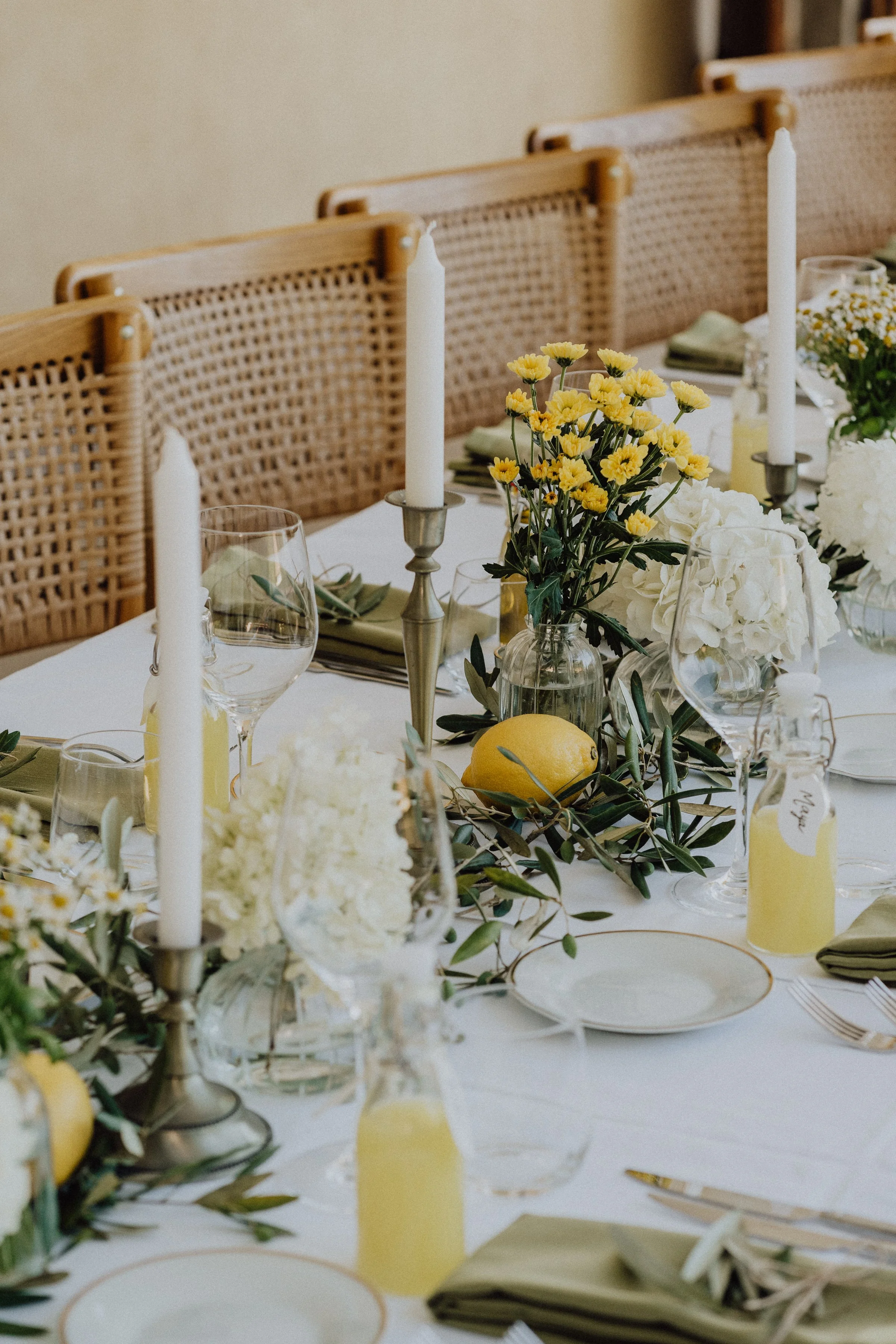 Une table de réception élégante et lumineuse, décorée dans un style méditerranéen. Des citrons frais, des branches d’olivier, et des fleurs jaunes et blanches apportent une touche de fraîcheur et de naturel. Les bougies longues dans des chandeliers d