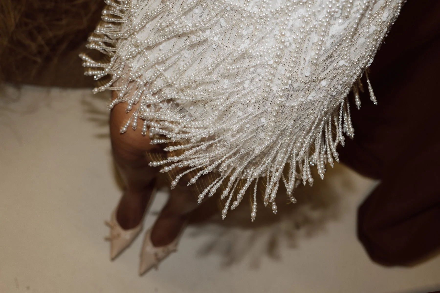 Détail des franges perlées d’une robe de mariée en mouvement au-dessus de chaussures à talons, photo de mariage au style éditorial chic et lumineux